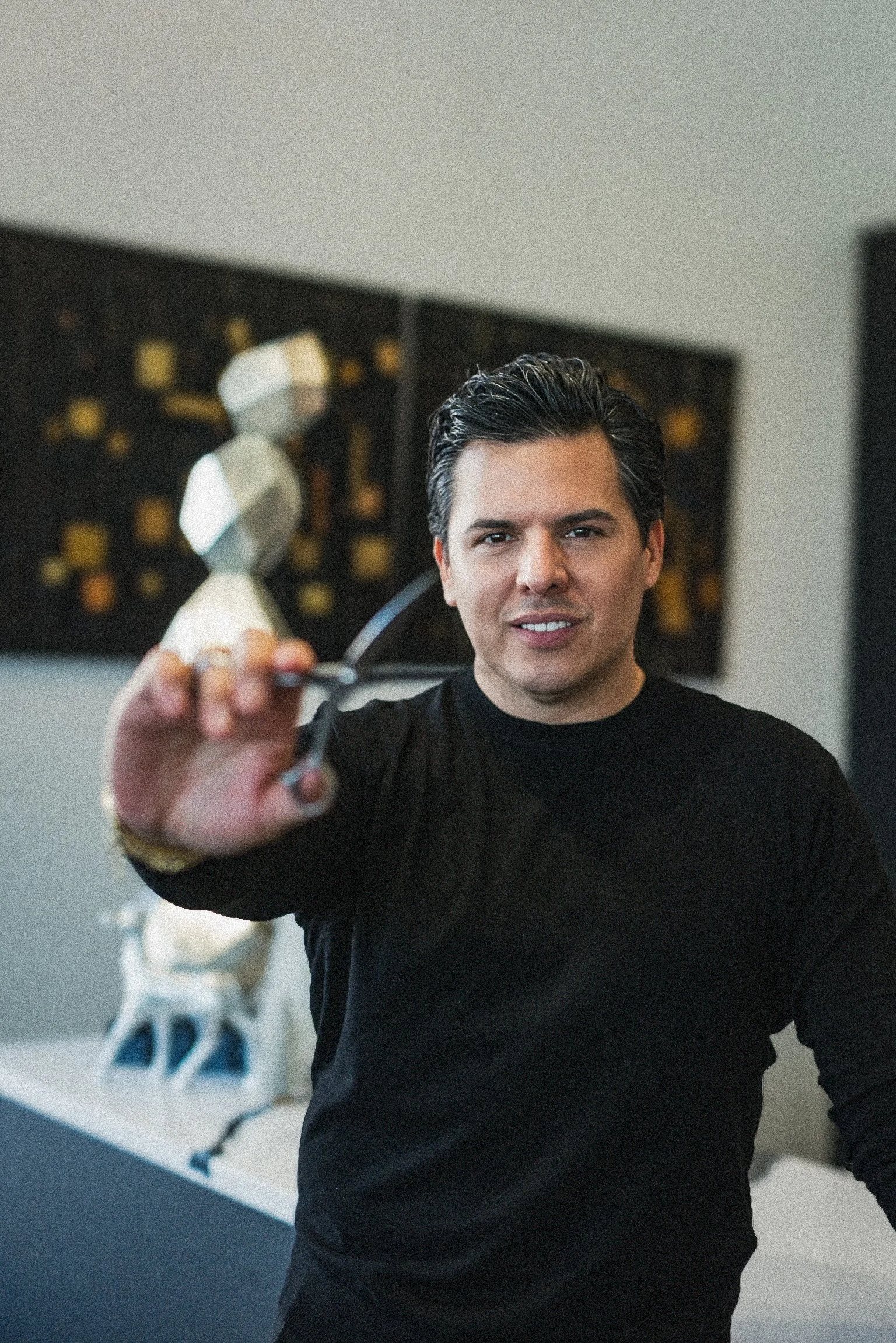 A man with dark hair and a black shirt holding a pair of scissors towards the camera in an indoor setting.