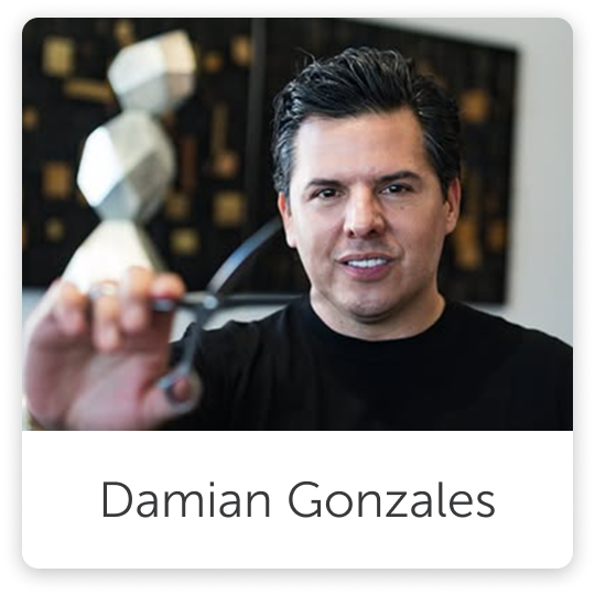 A man with dark hair smiling and holding glasses in front of a camera, with a blurred background and a name tag that reads Damian Gonzales.