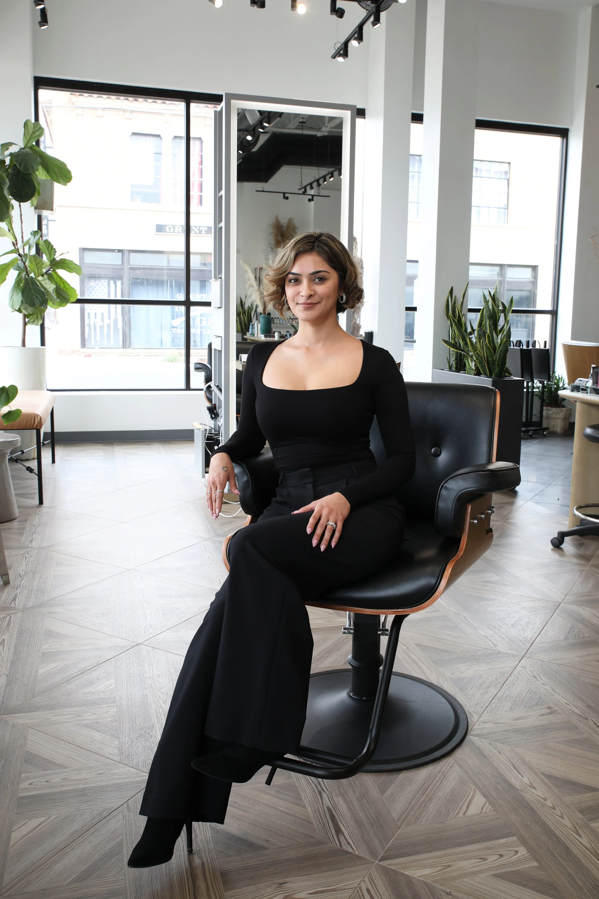 A woman sitting and posing in a salon chair wearing black clothes in an indoor setting