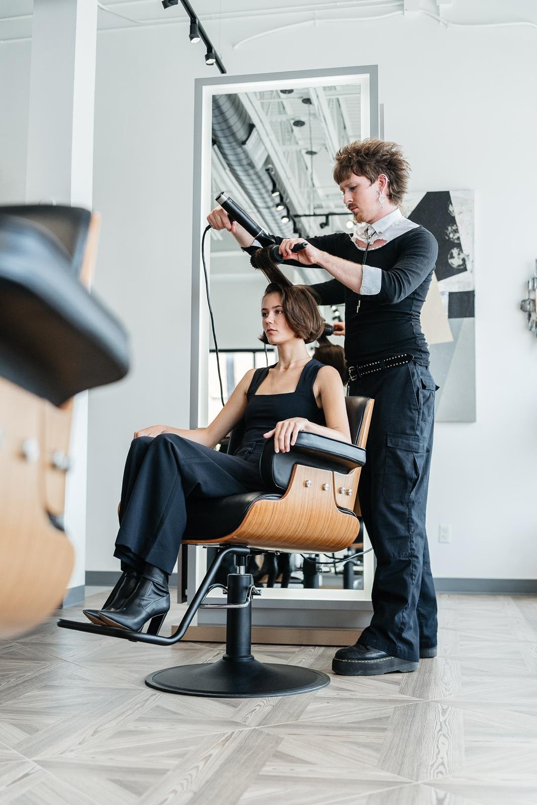 A hairstylist blow-drying a woman's hair in a modern salon with a large mirror.