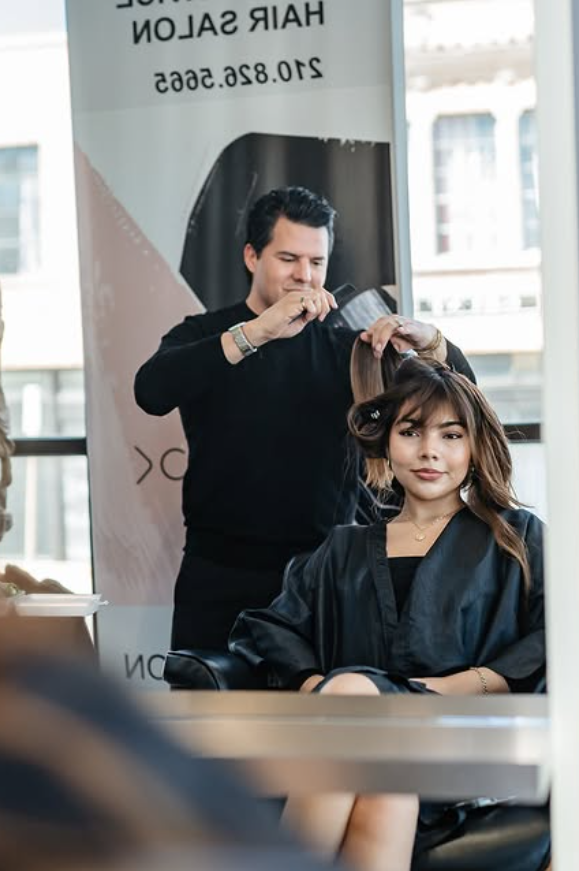 A hairstylist blow-drying a woman's hair in a salon with large windows and a city view in the background.