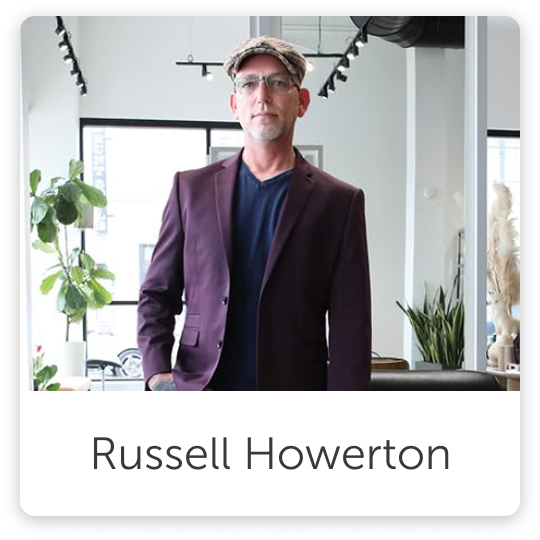 A man wearing a purple blazer, dark shirt, glasses, and a flat cap standing indoors with green plants and large windows in the background.