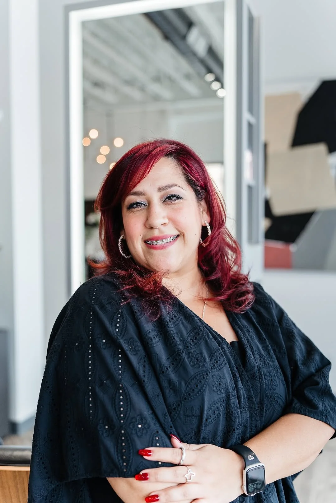 A woman with red hair, wearing a black embroidered top, hoop earrings, and a smartwatch, smiling with arms crossed in a modern indoor setting.