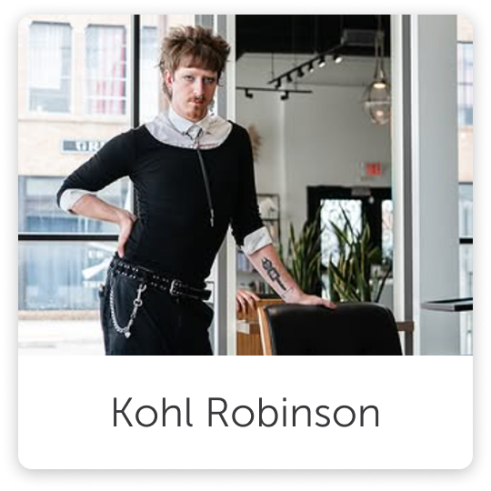 A man with light brown, tousled hair and a mustache stands in a modern, well-lit room with large windows, wearing a black sweater with a collared shirt underneath, and black pants with chain accessories.