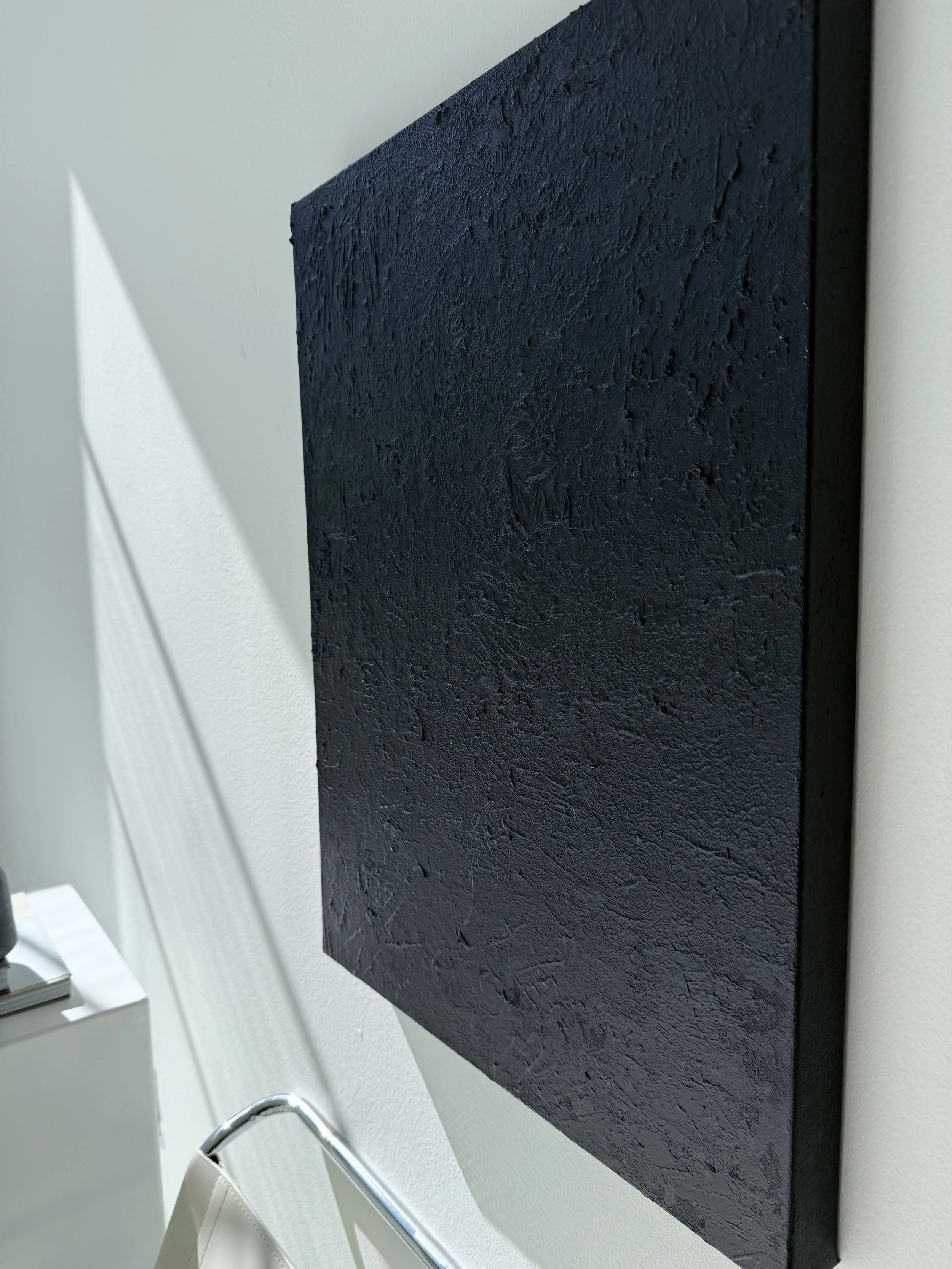 A black textured canvas artwork mounted on a white wall, with shadows cast on the left side.