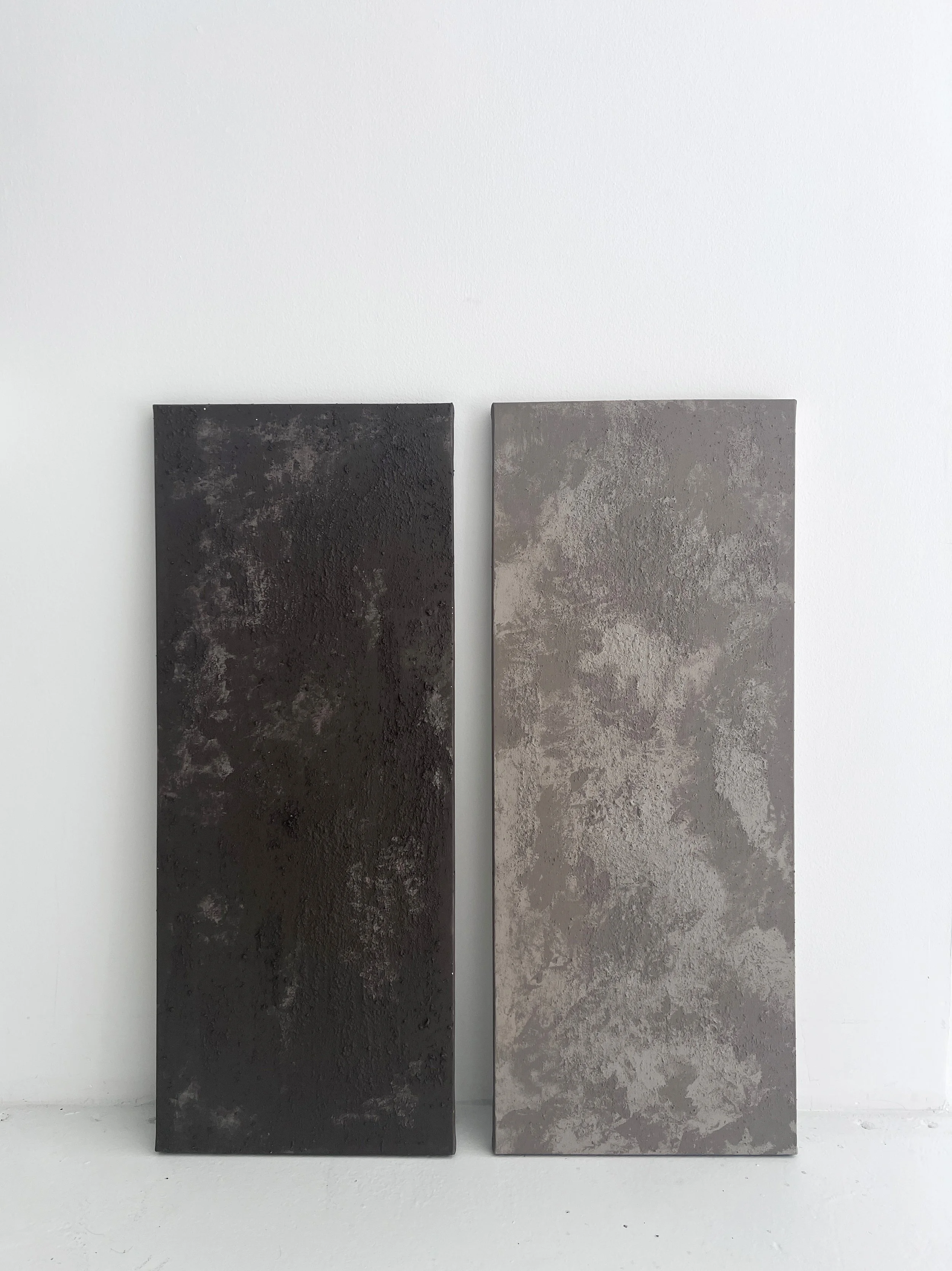 Two rectangular textured concrete paintings, one dark brown and the other light gray, leaning against a white wall.