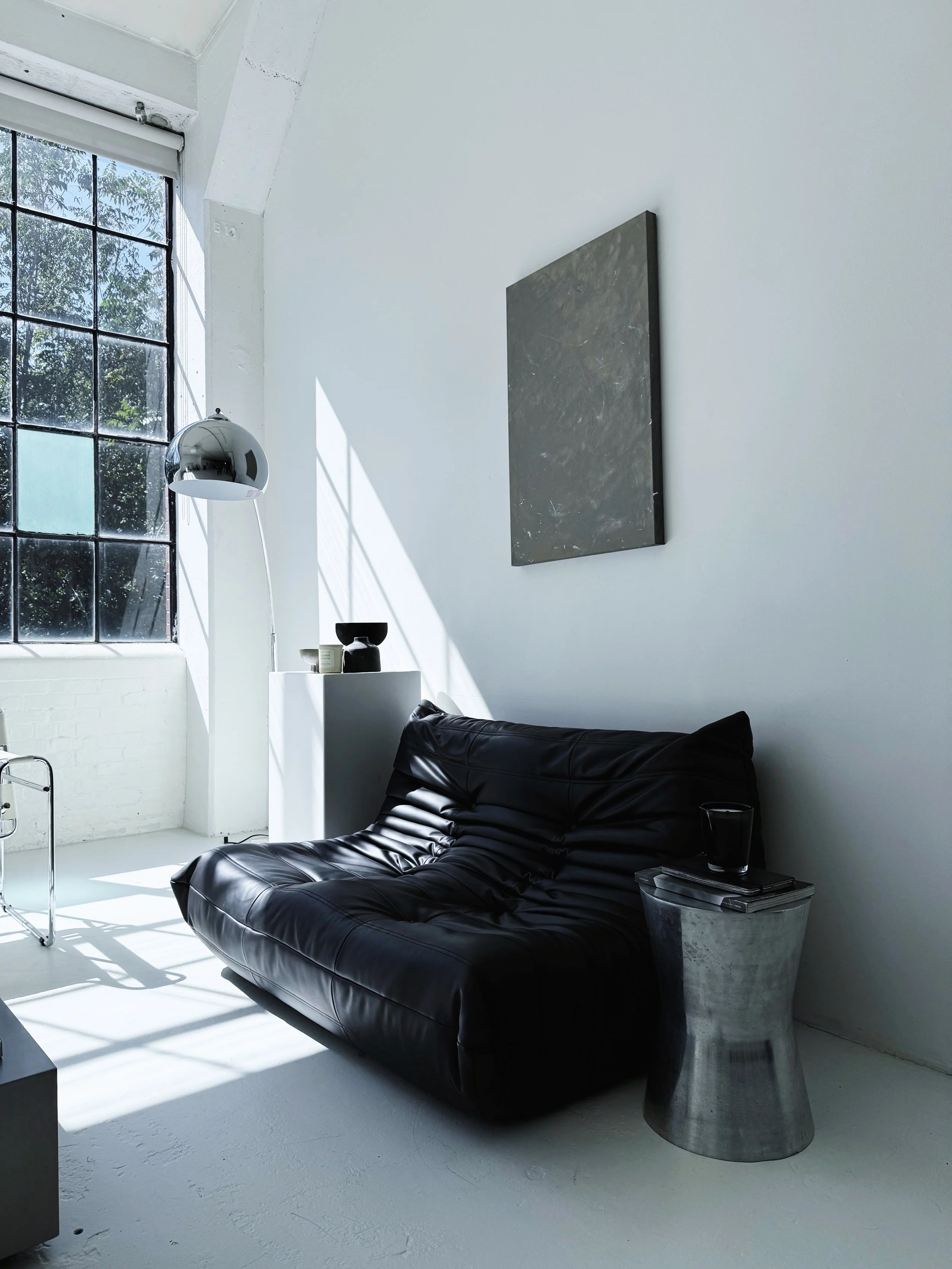 Modern living room with a black leather sofa, a metal side table with a glass, a white pedestal with black and white decorative objects, and a large concrete inspired abstract painting mounted on the wall