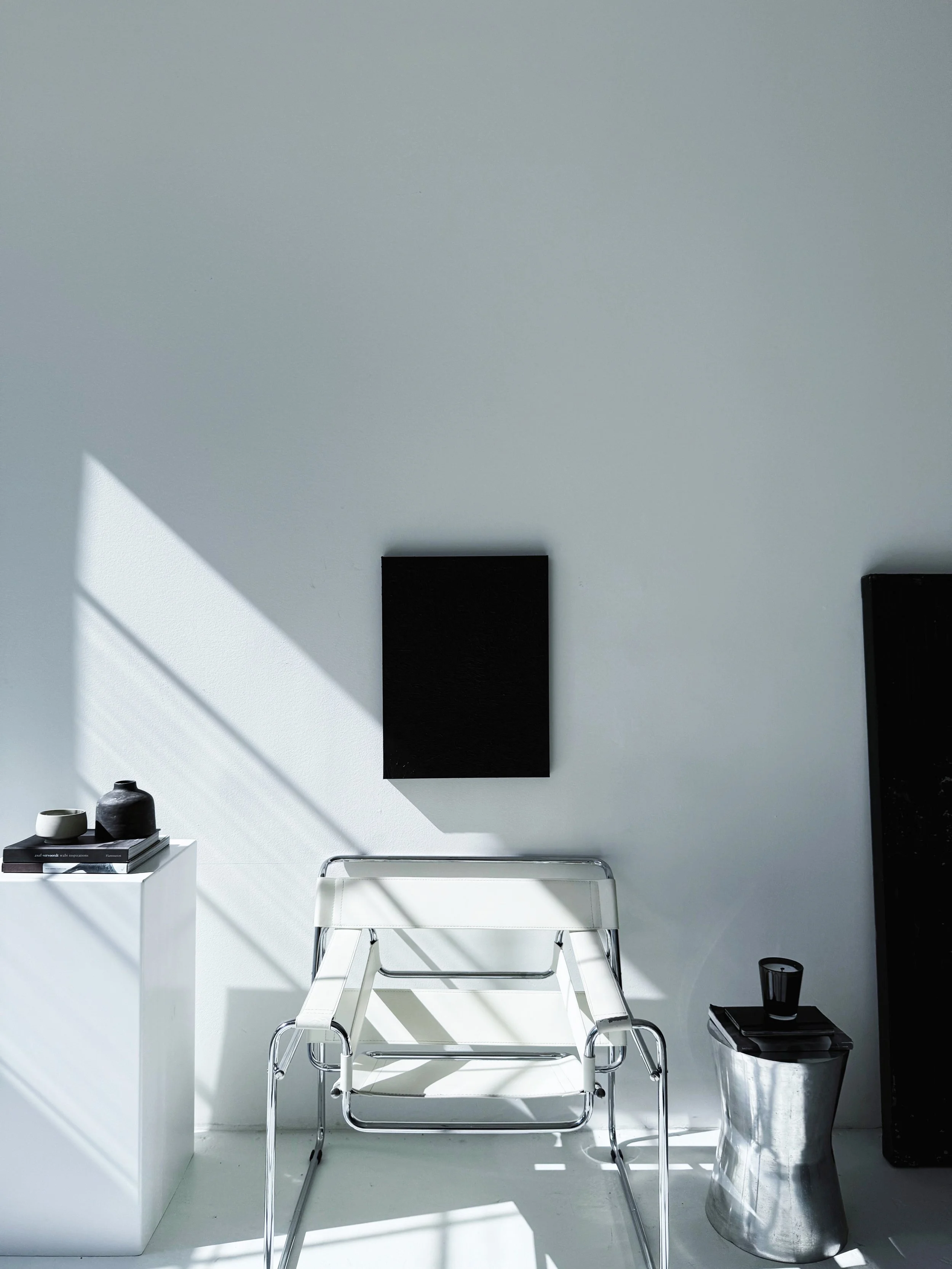 Minimalist white armchair with chrome frame in a room with white walls and floor, black wall art, black tray with a black candle, black vase, and white and black decorative objects.