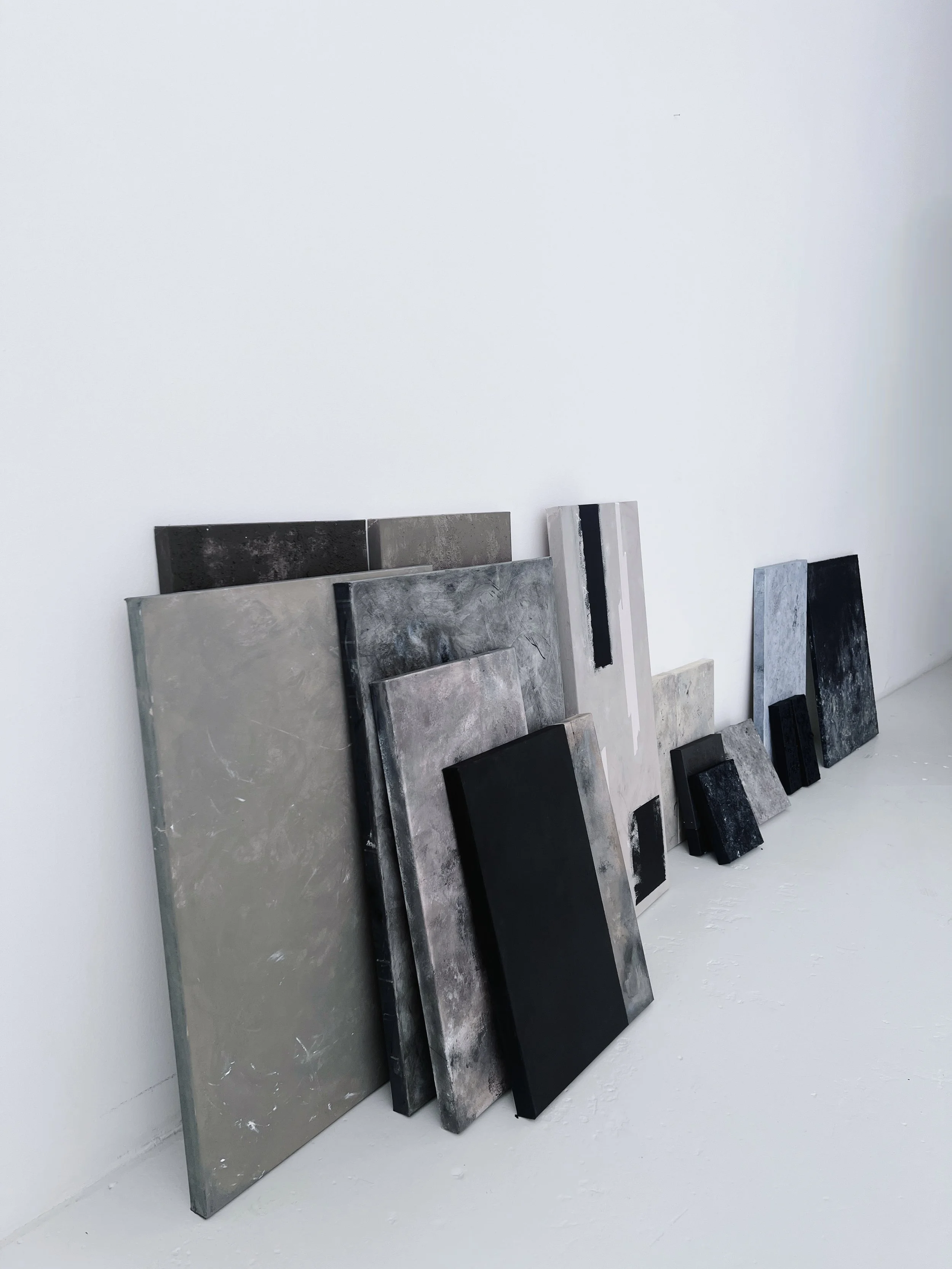 An arrangement of various-sized black, gray, and white rectangular abstract paintings leaning against a white wall.