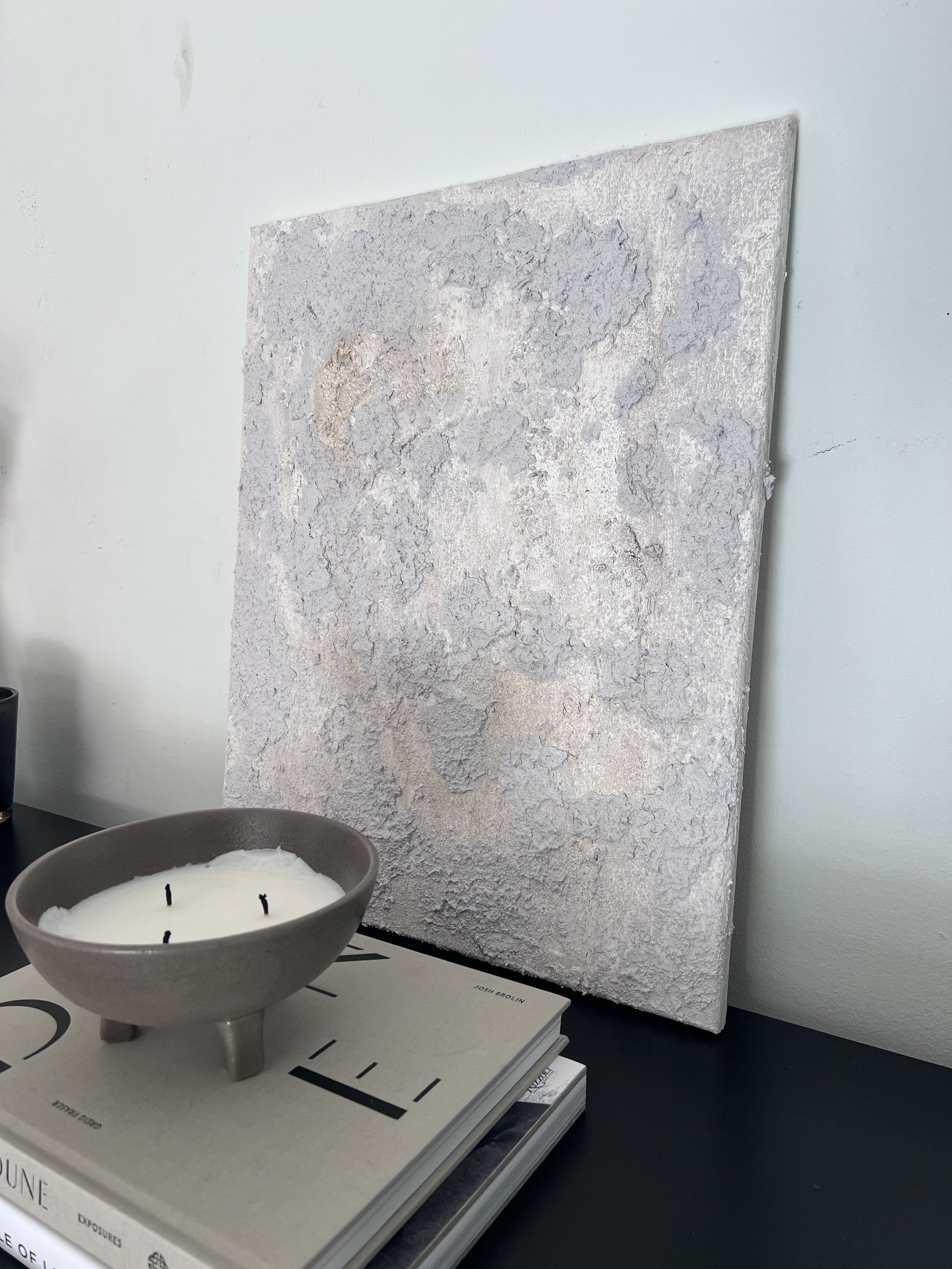 A textured white canvas leaning against a white wall, with a gray bowl containing a white candle with three wicks placed on a stack of books on a black surface.