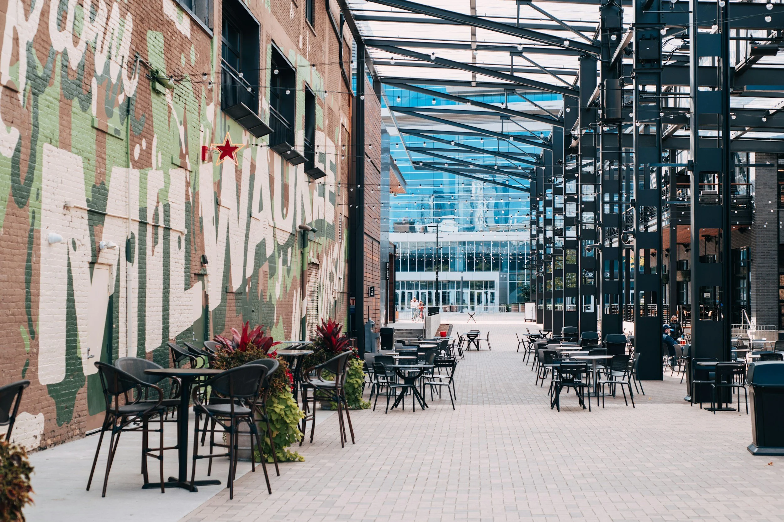 Outdoor seating area with black tables and chairs in front of a brick wall painted with green, white, and brown camouflage and the words 'The Wau' visible. String lights hang overhead, and a modern glass building is in the background.