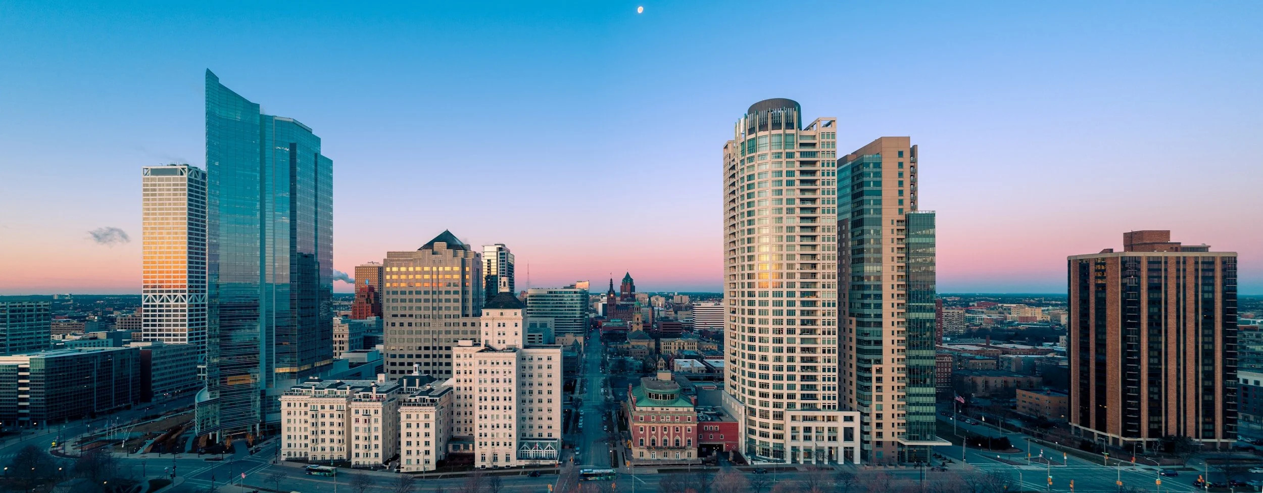 City skyline at sunset with modern high-rise buildings and a visible moon in the sky.
