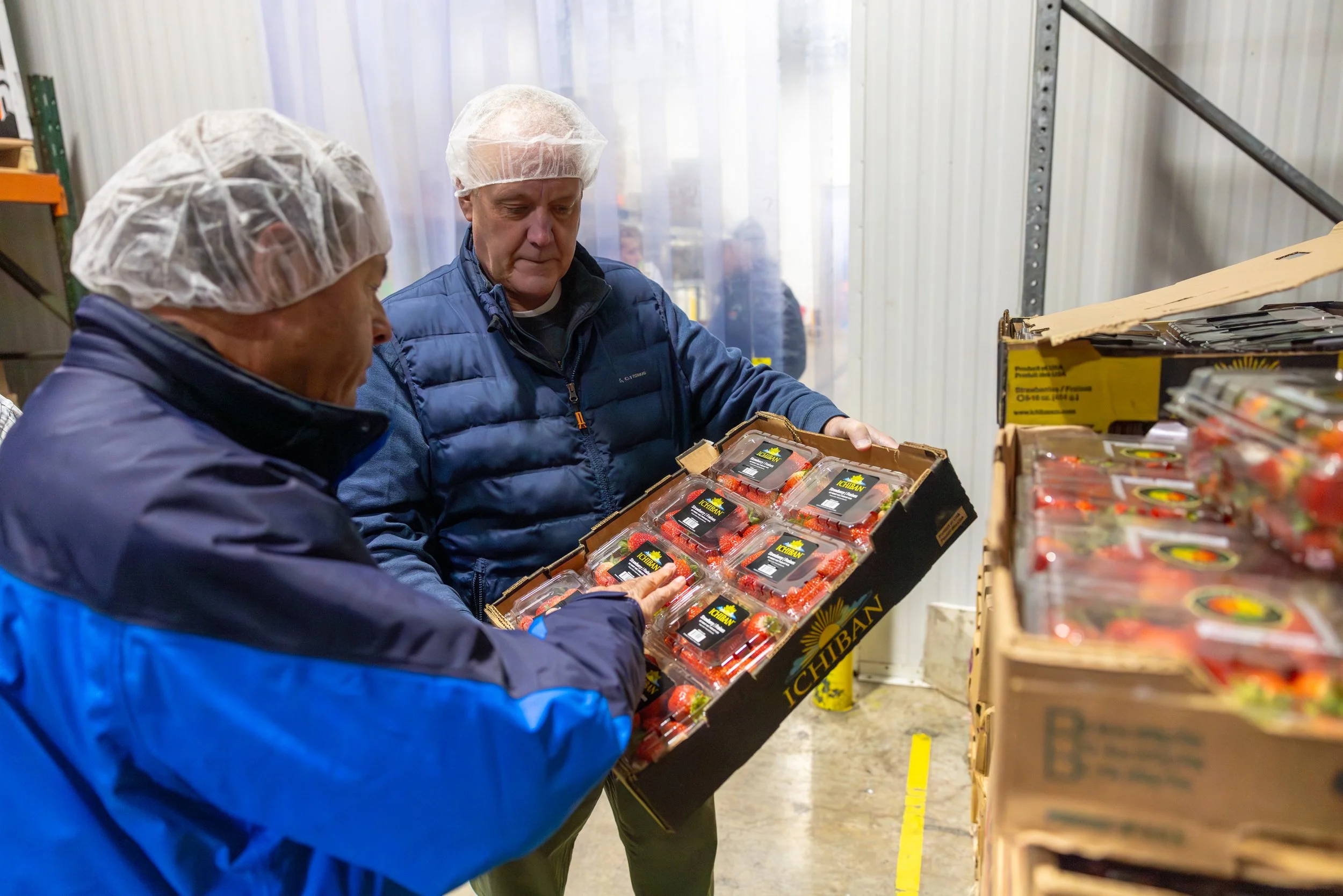 Two men wearing hairnets and jackets inspecting a box of strawberries at a warehouse.