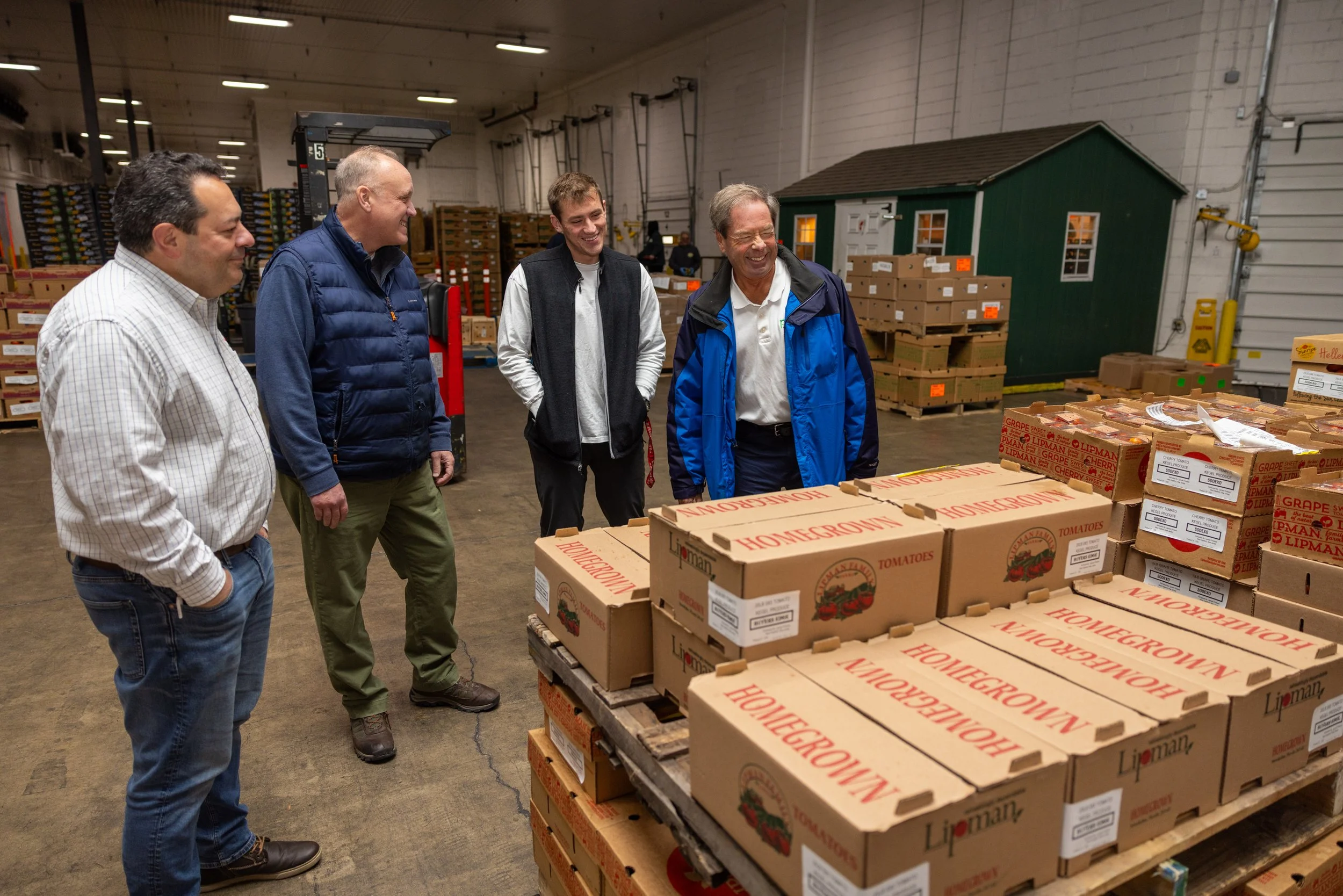 Four men are standing in a warehouse near boxes of Tomatoes, some of which are labeled Homegrown. They are smiling and looking at the boxes.