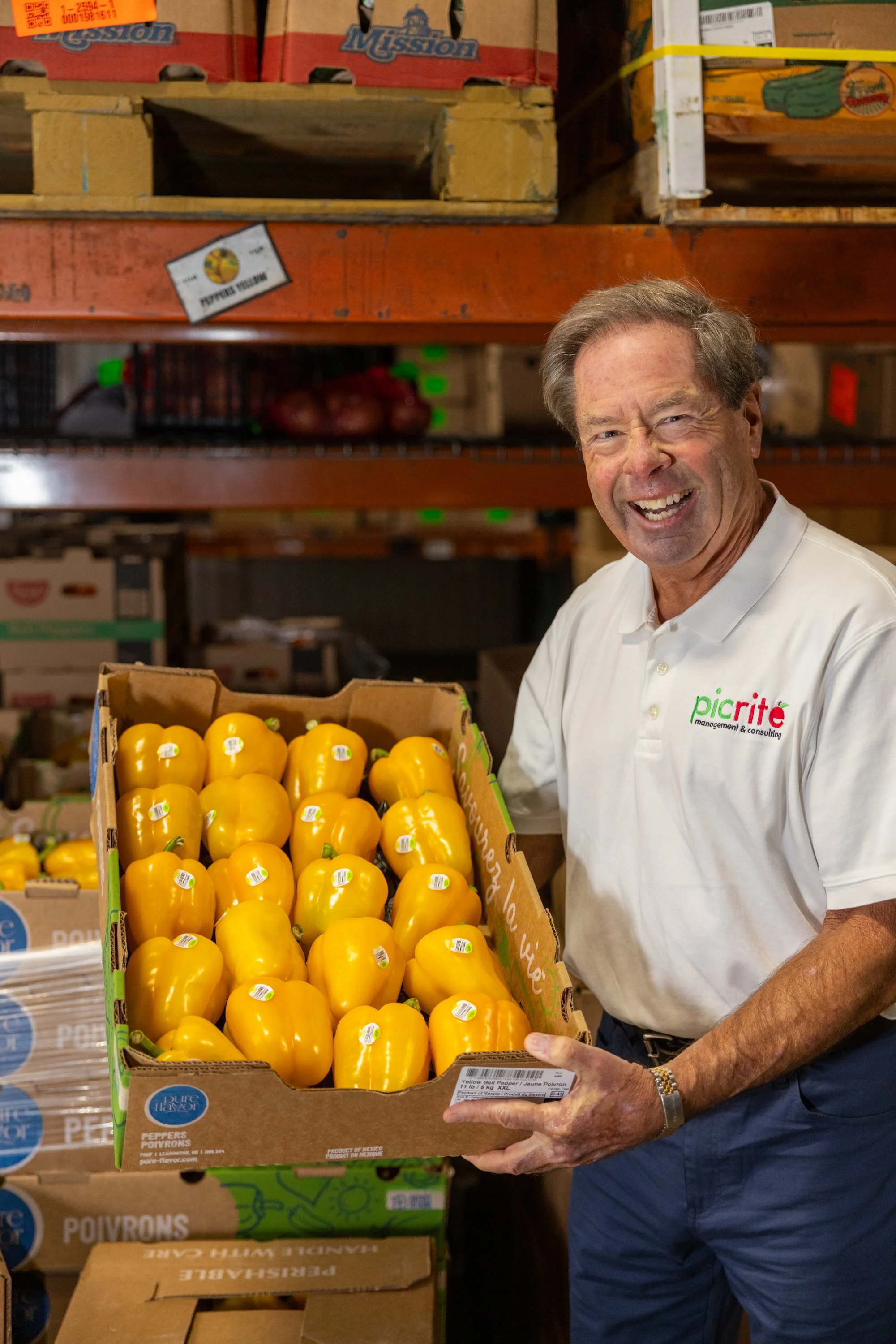 A man smiling and holding a box of yellow bell peppers in a warehouse or grocery storage area.