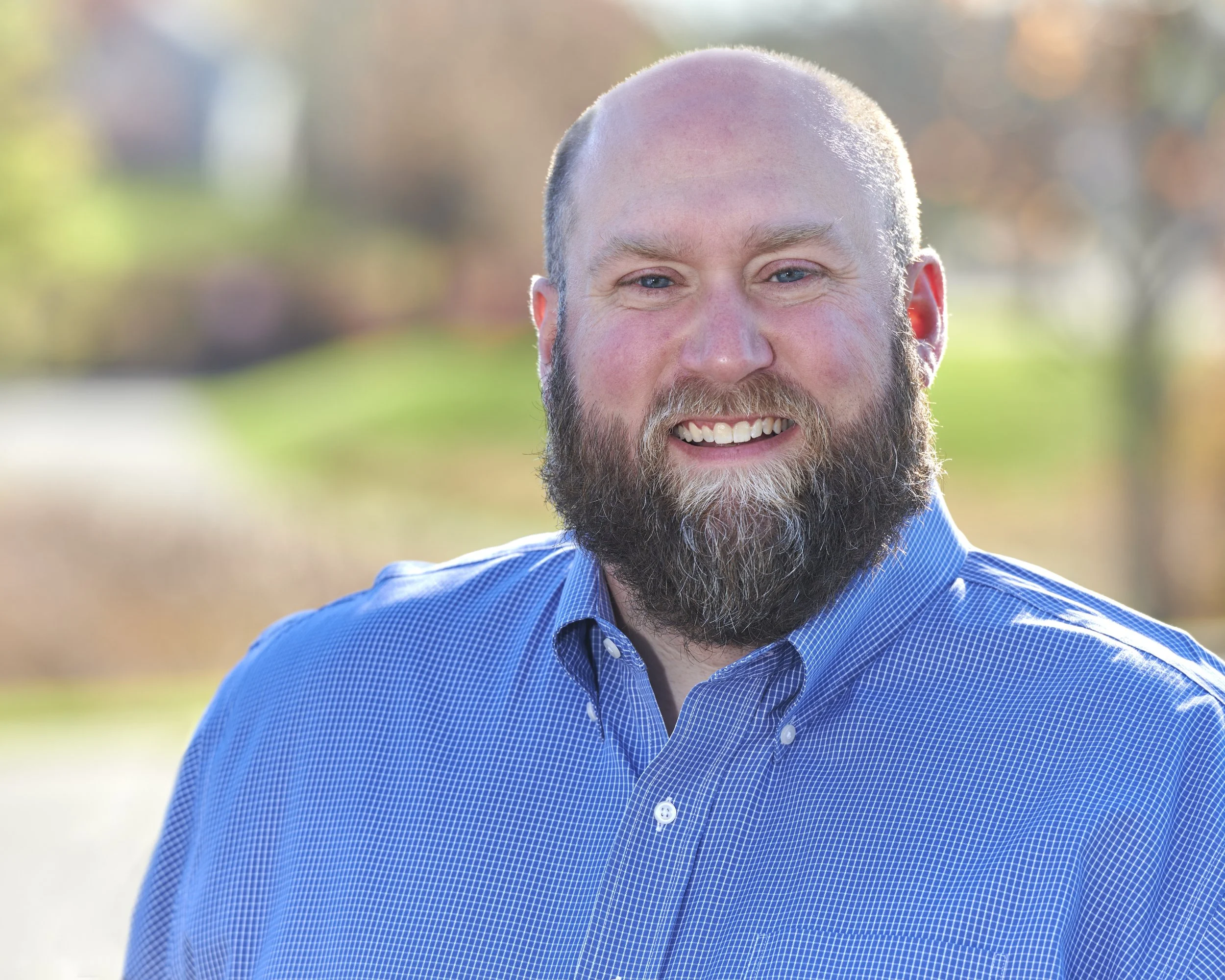 A smiling man with a beard and a bald head, wearing a blue checkered shirt, outdoors with a blurred background of trees and sunlight.