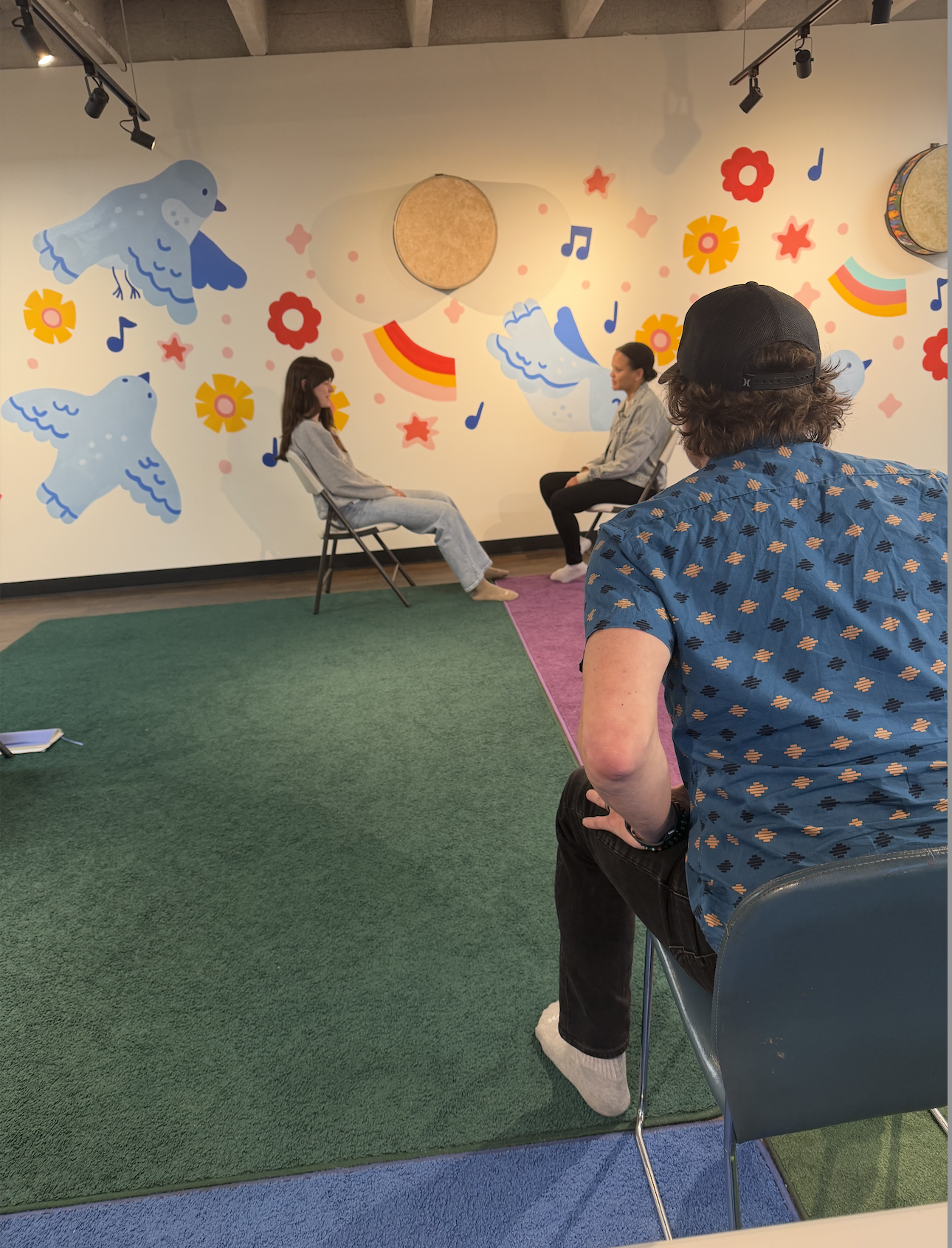 A woman and a girl are seated and facing each other in a room with colorful, cartoon-style illustrations of birds, flowers, rainbows, and musical notes on the wall behind them. A person with a black cap and patterned blue shirt is sitting with their back to the camera, observing the scene.