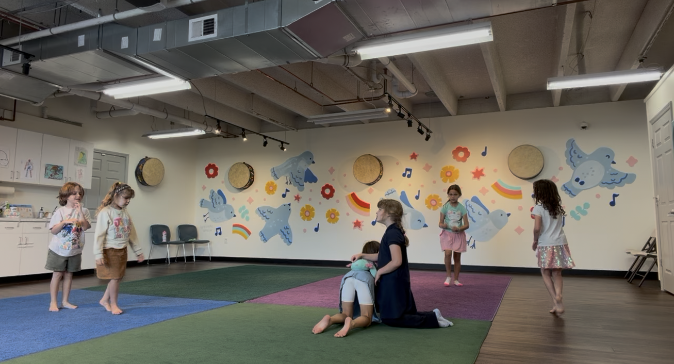 Children playing in a room with colorful wall decorations of birds, flowers, rainbows, and music notes. Two girls are hugging on the floor, while three others stand nearby.