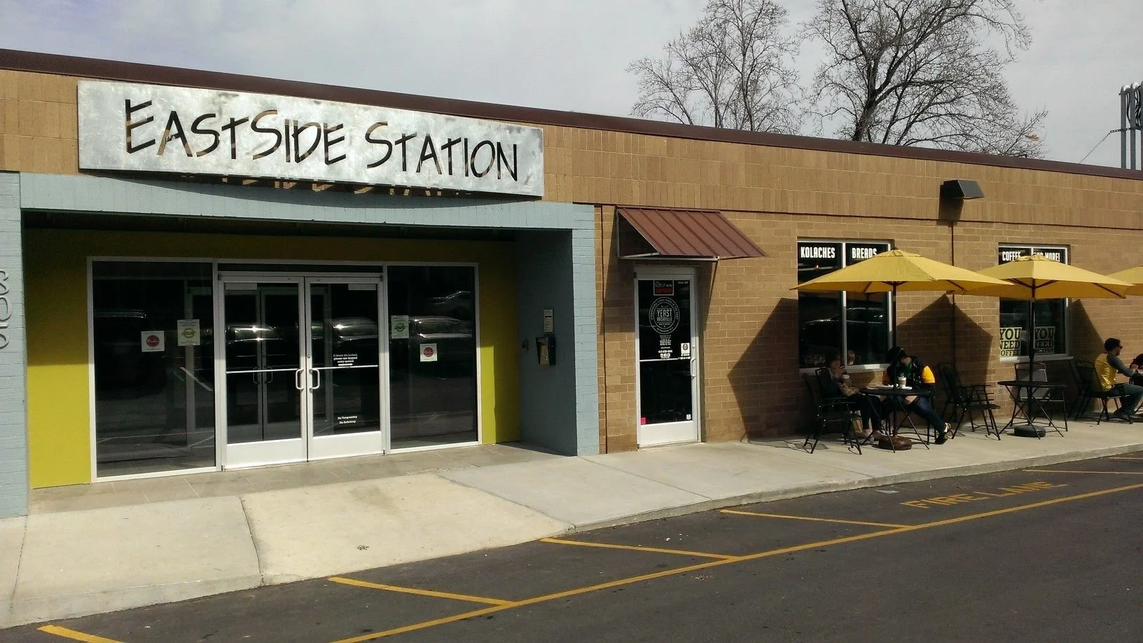 Exterior of Eastside Station with a sidewalk, parked cars, and outdoor seating with yellow umbrellas, people sitting outside, and a brick building with signage.