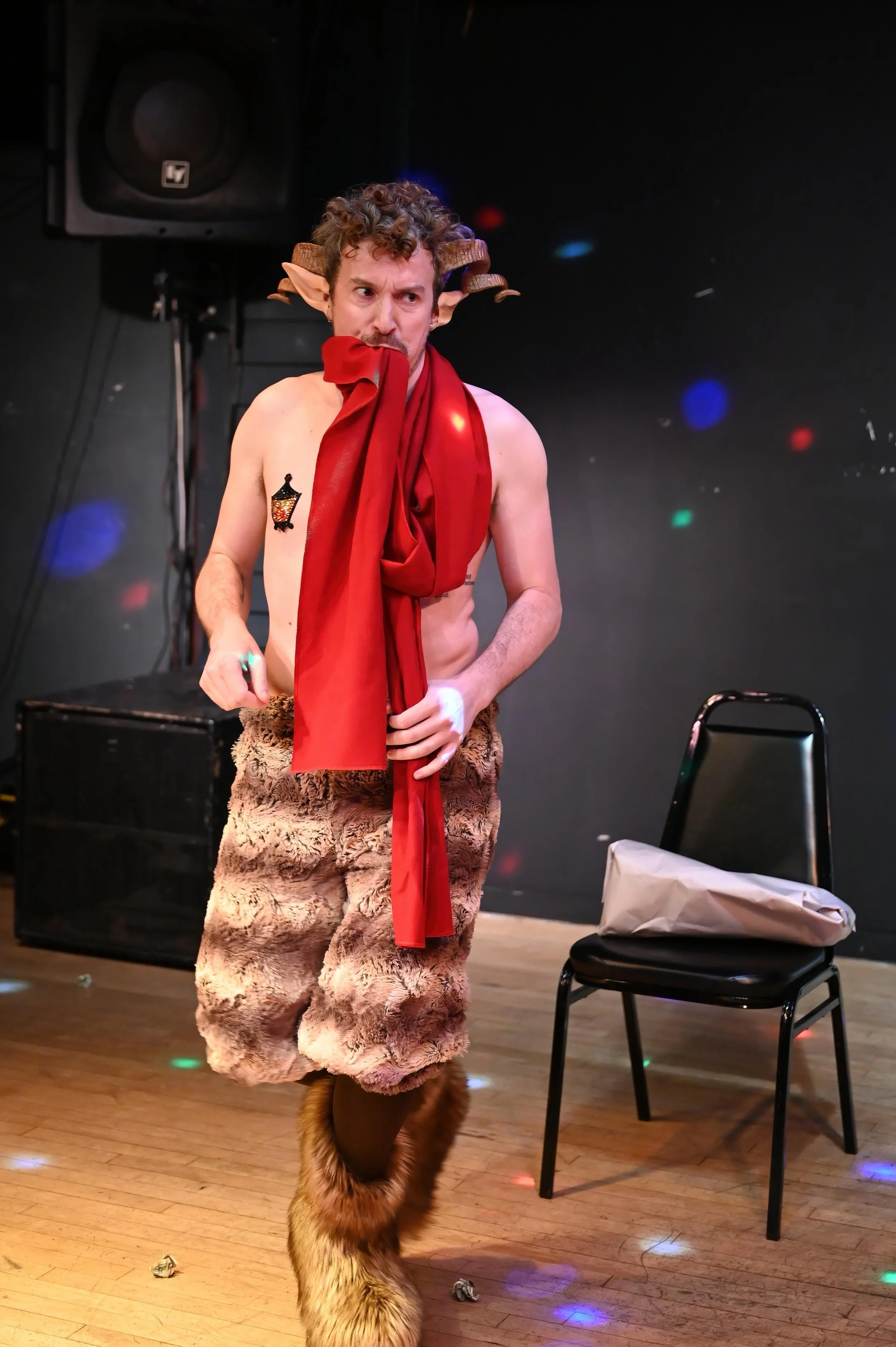 A man dressed in costume, resembling a mythical creature with curly hair, headphones, a red scarf, fur pants, and furry boots, stands on a stage with a black background and multicolored lights.