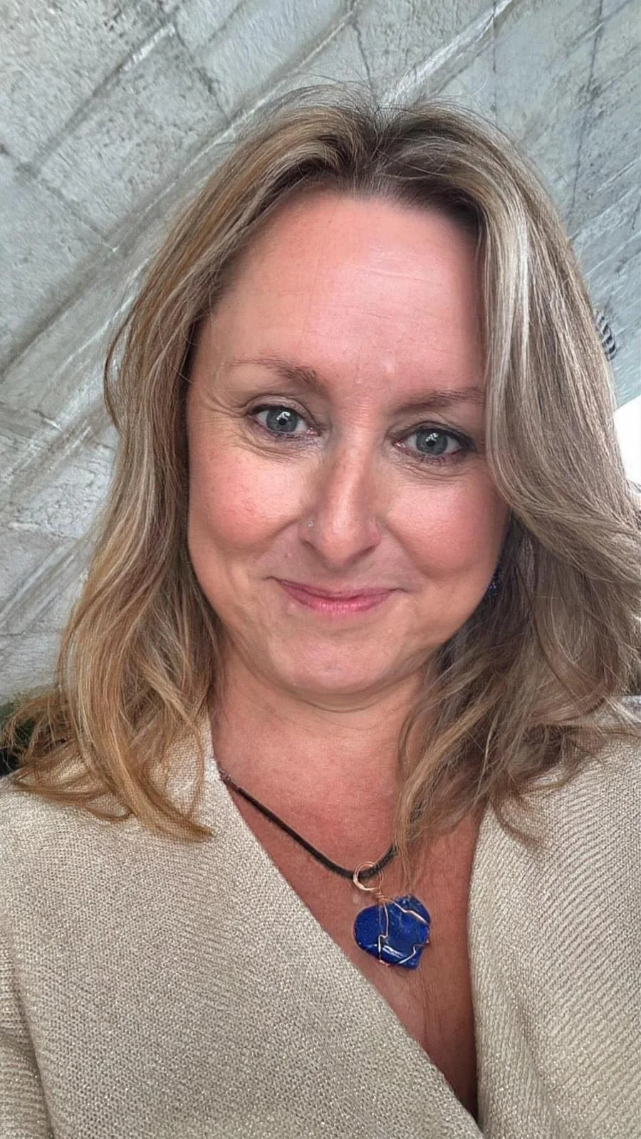 A woman with shoulder-length blonde hair and blue eyes smiling, wearing a beige top and a blue gemstone pendant necklace, standing against a stone wall background.