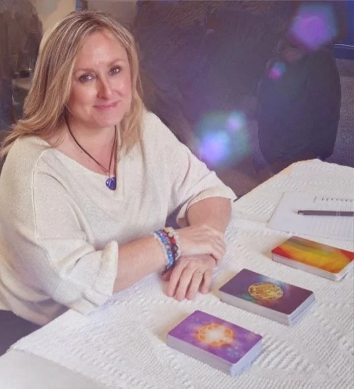 A woman sitting at a table with tarot cards, a notebook, and a pen, wearing a white sweater and colorful bracelets.
