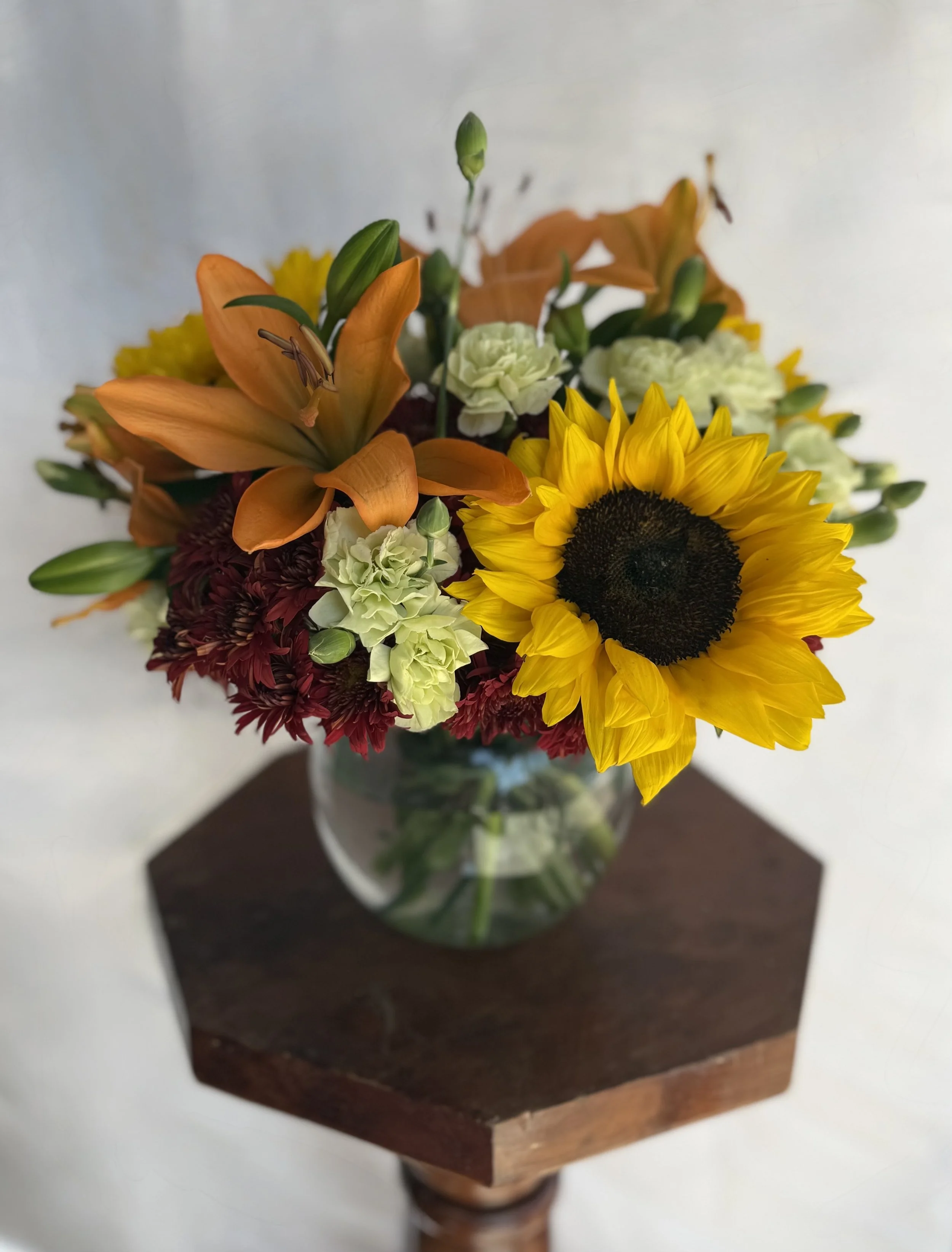 A glass vase filled with a colorful bouquet of flowers, including orange lilies, yellow sunflowers, white carnations, and red chrysanthemums, sitting on a small wooden table against a white background.