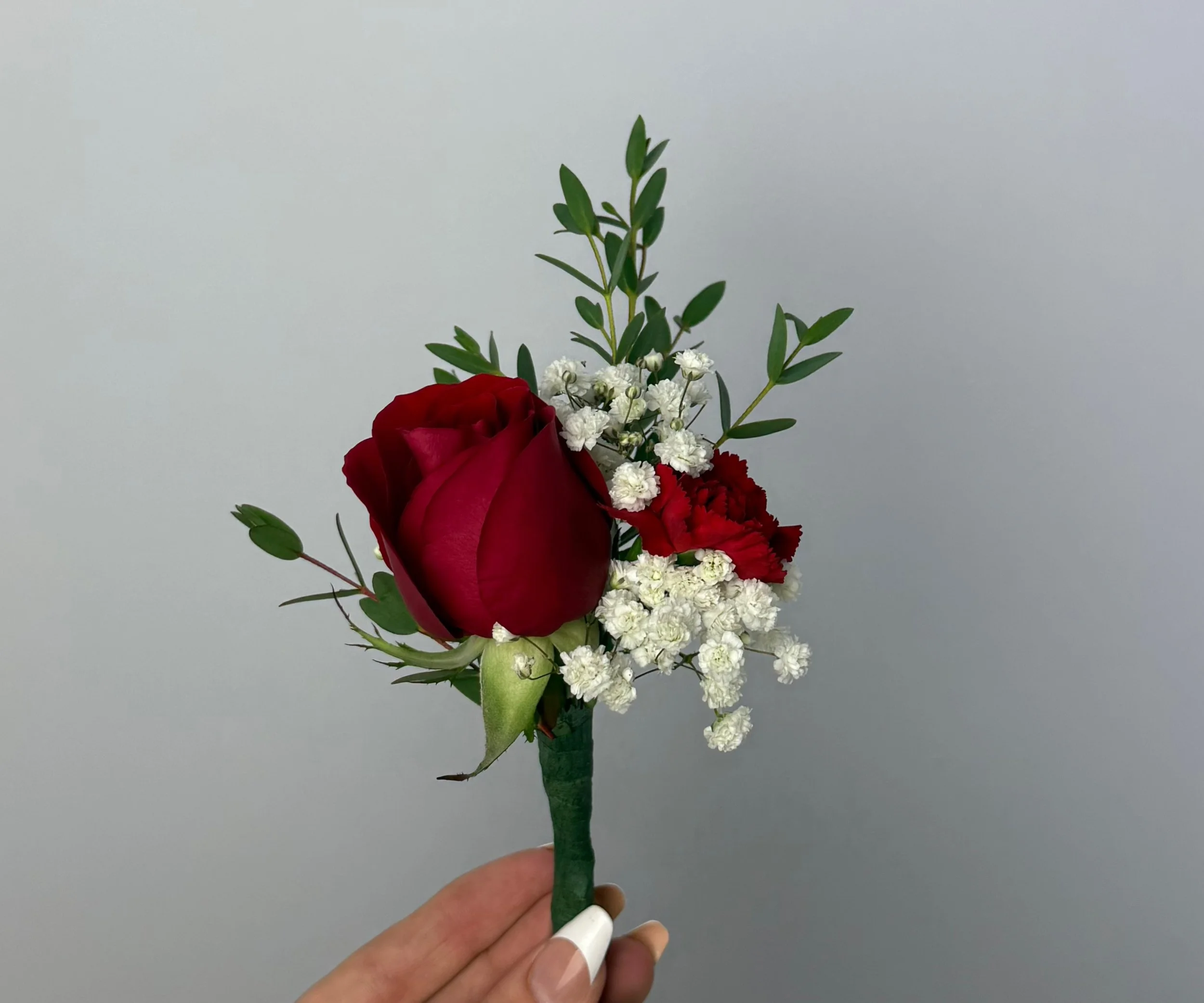 Hand holding a boutonniere with a red rose, white baby's breath, red carnations, and green leaves against a plain background.