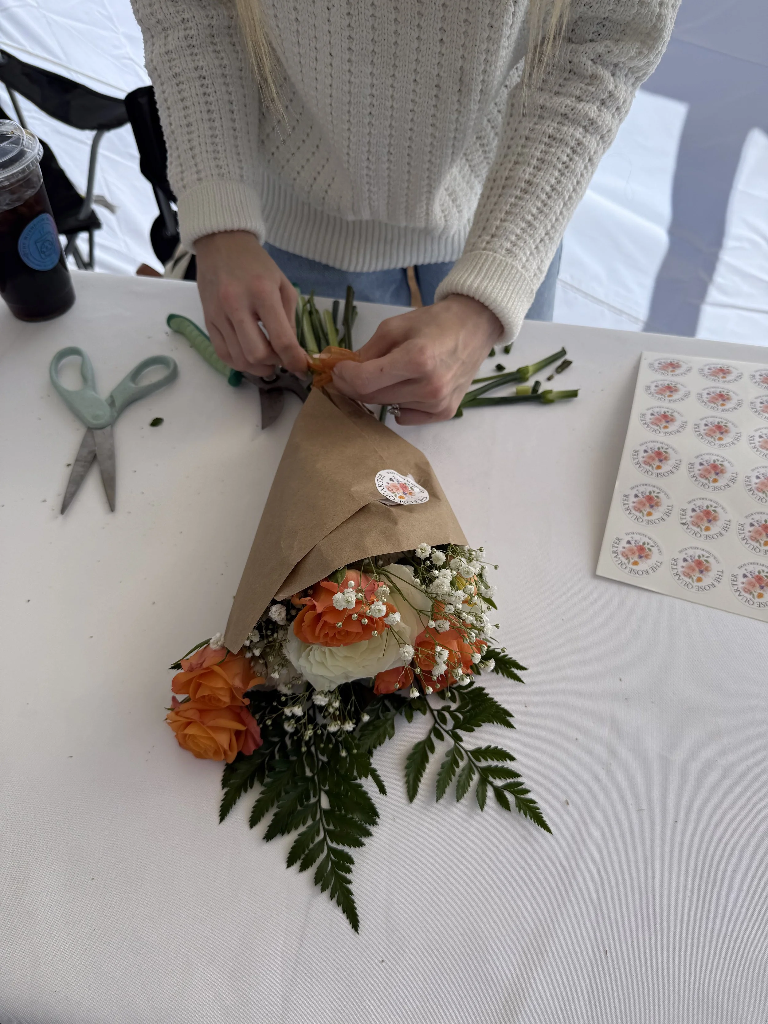 Person wrapping a bouquet of orange roses and white flowers in brown paper on a white table, with open scissors, a sticker sheet, and a drink nearby.