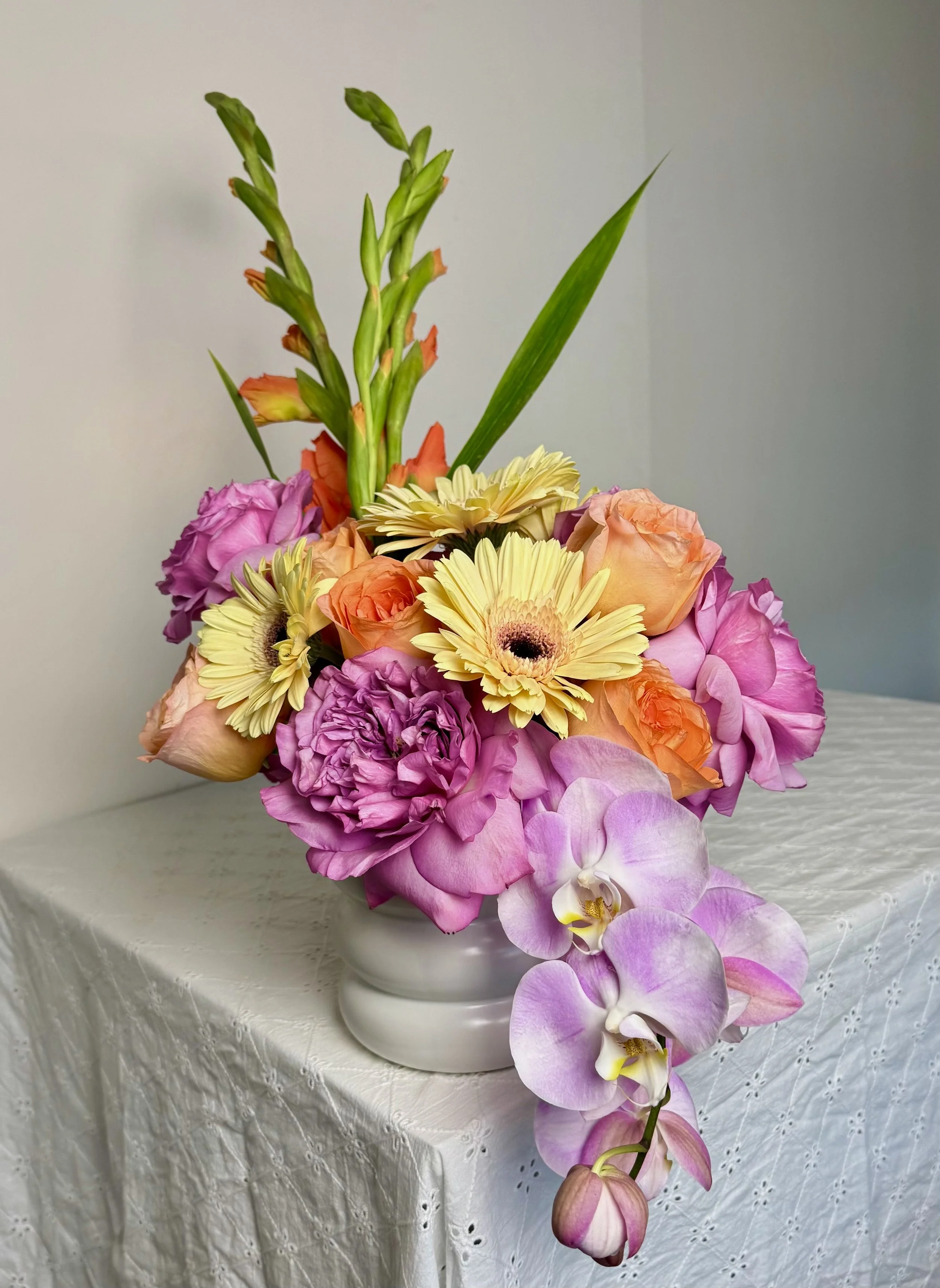 Colorful flower arrangement with pink roses, yellow gerbera daisies, orange gladiolus, purple peonies, and pink orchids in a silver vase on a white lace tablecloth against a light-colored wall.
