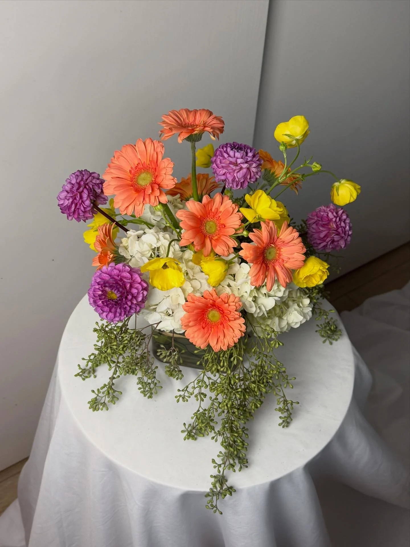 Colorful flower arrangement with pink gerbera daisies, purple chrysanthemums, yellow ranunculus, white hydrangeas, and green foliage in a black vase on a white round table.