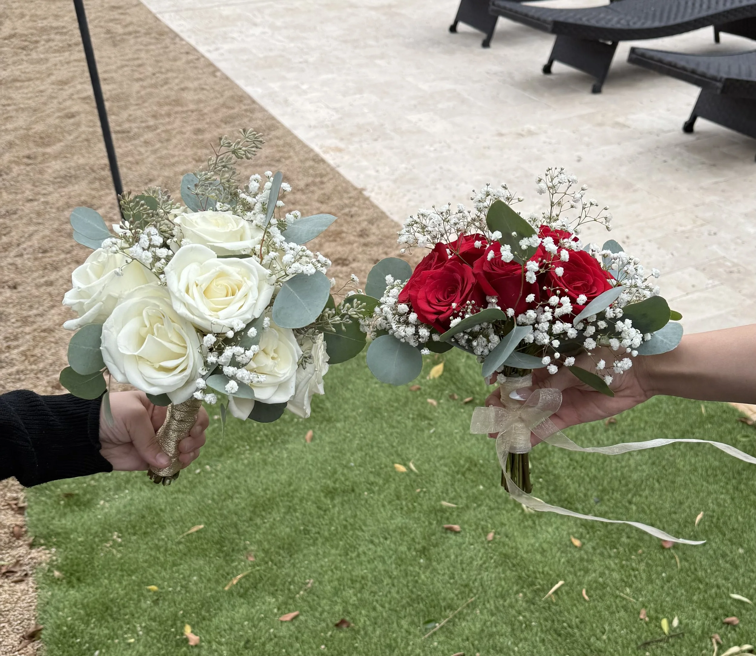 Two people each holding a bouquet of flowers outside on grass and pavement. One bouquet has white roses and eucalyptus, the other has red roses and baby's breath.