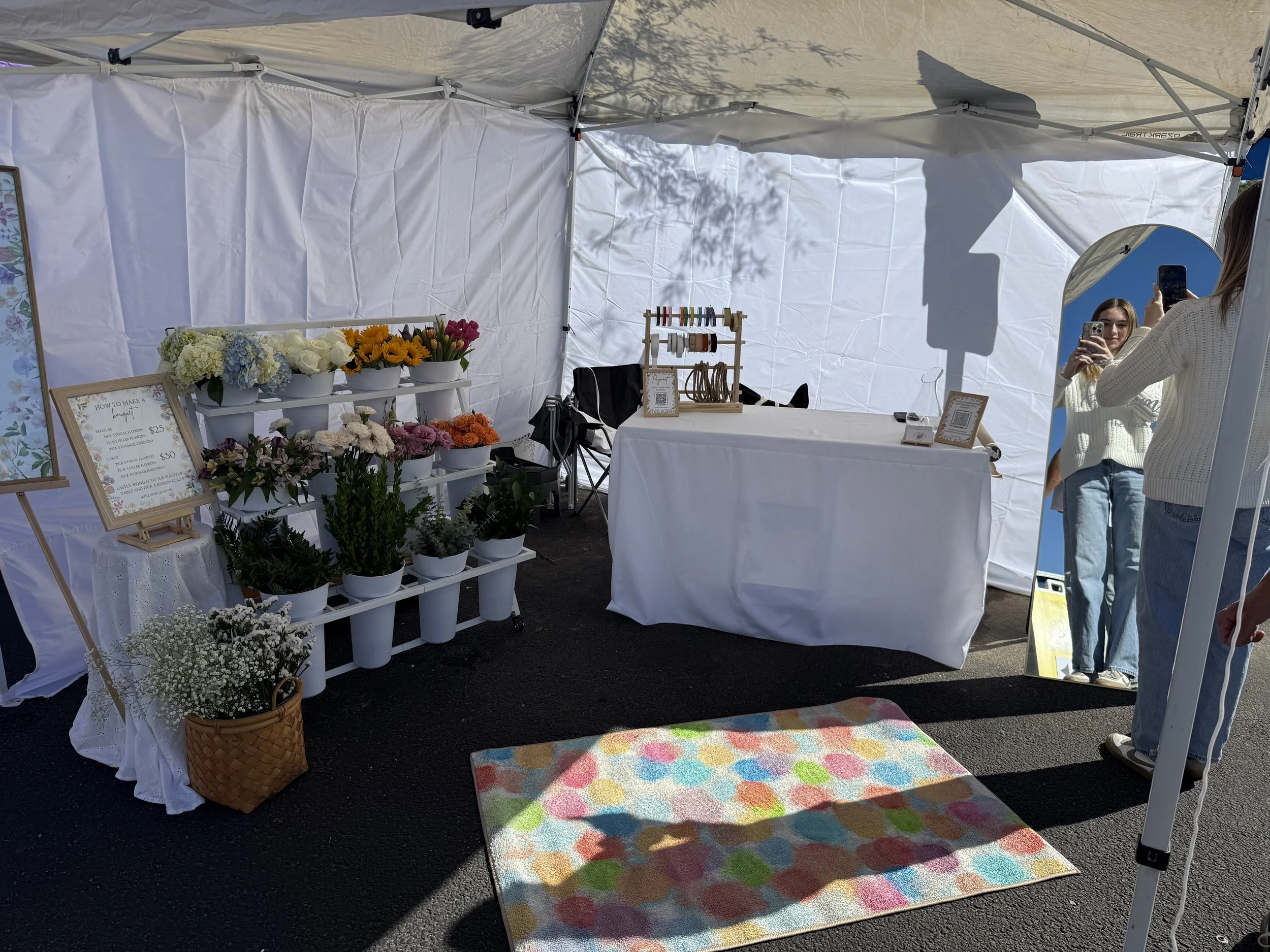 A flower and craft booth under a white canopy tent, with various flower arrangements, a display of thread spools, and a mirror, with two women taking photos.