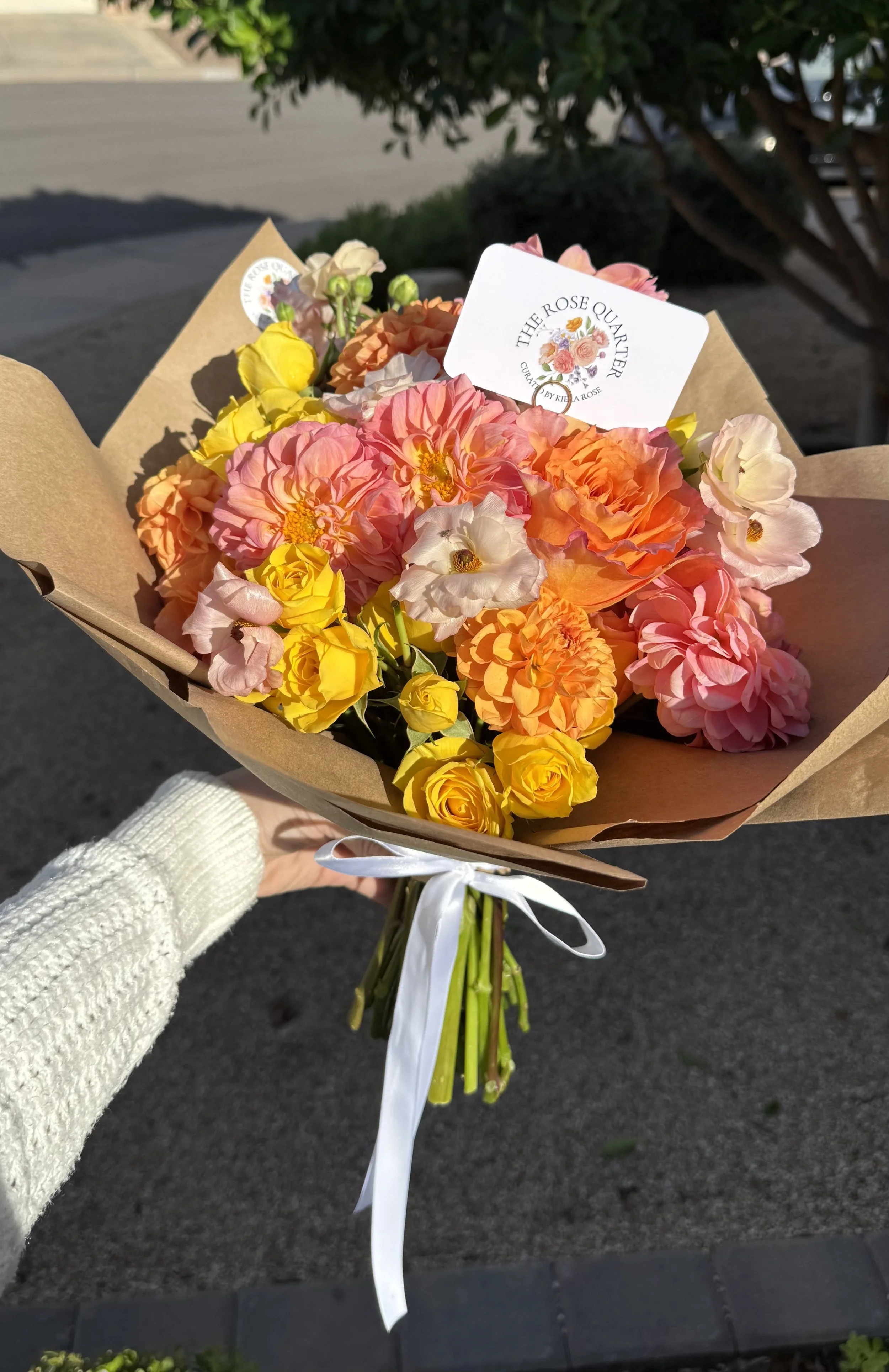 A person wearing a white sweater holding a bouquet of mixed pink, yellow, and white flowers wrapped in brown paper with a white ribbon, outdoors with sunlight and greenery in the background.