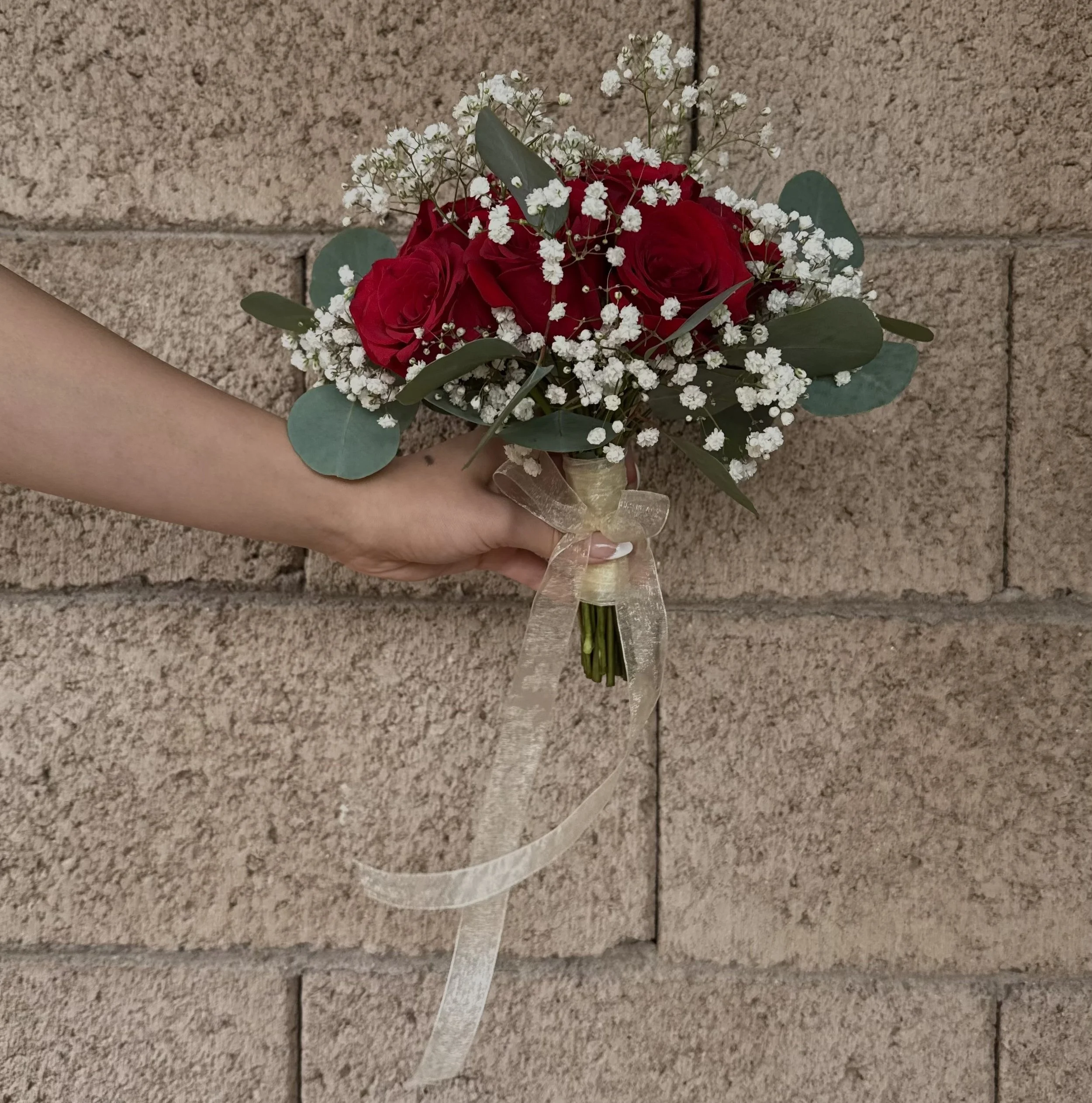 A hand holding a bouquet with red roses, white baby's breath, and eucalyptus leaves, wrapped with a light-colored ribbon against a brick wall.