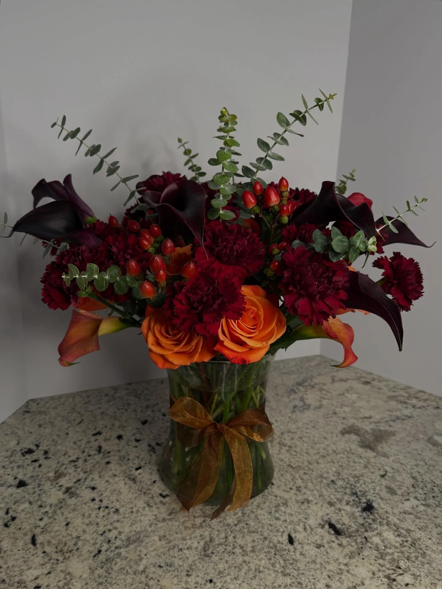 A bouquet of deep red, orange, and burgundy flowers in a glass vase with a brown ribbon, placed on a marble surface.