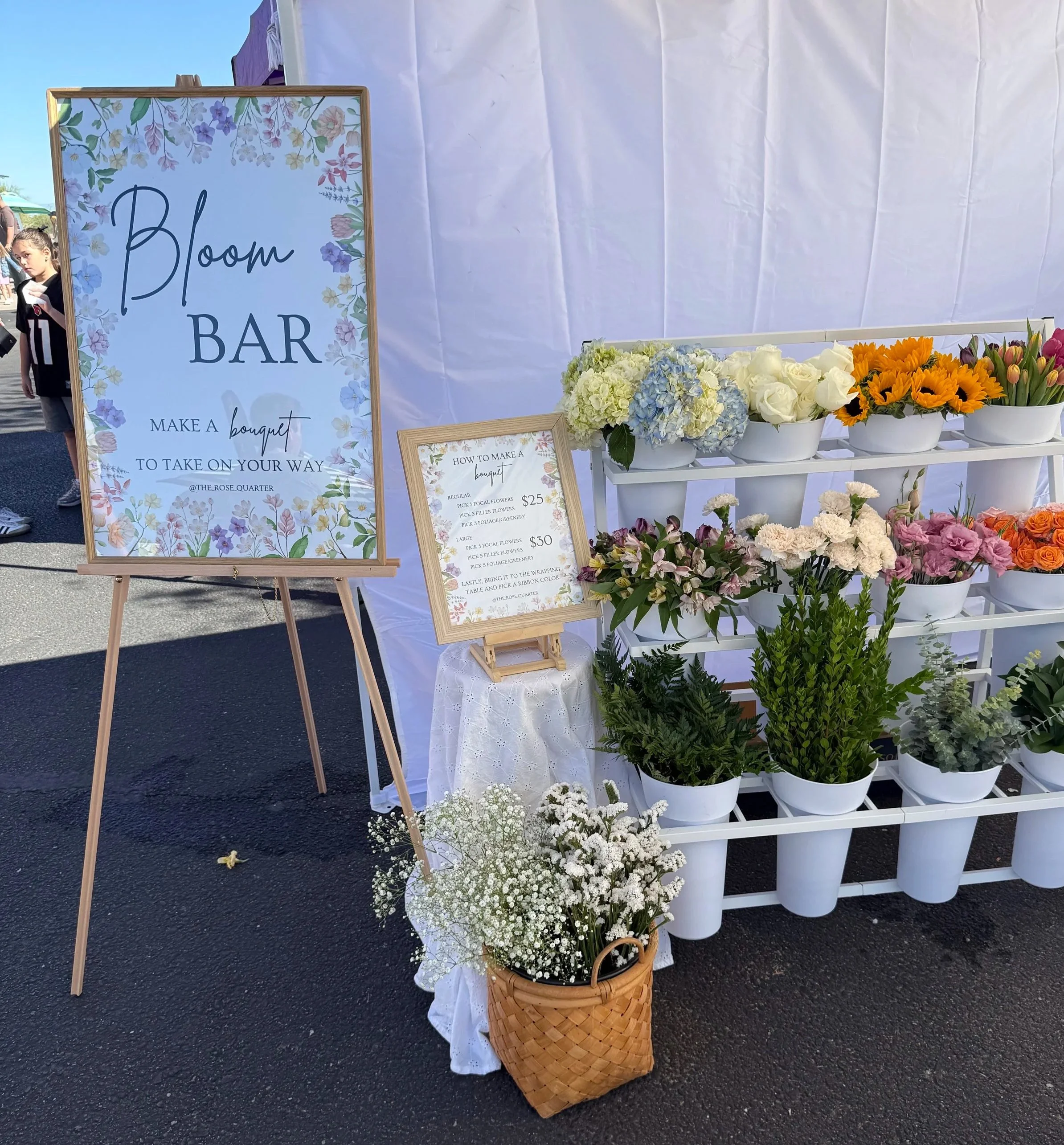 Flower display at a bloom bar with signs about making bouquets, potted flowers, and a woven basket filled with white baby's breath