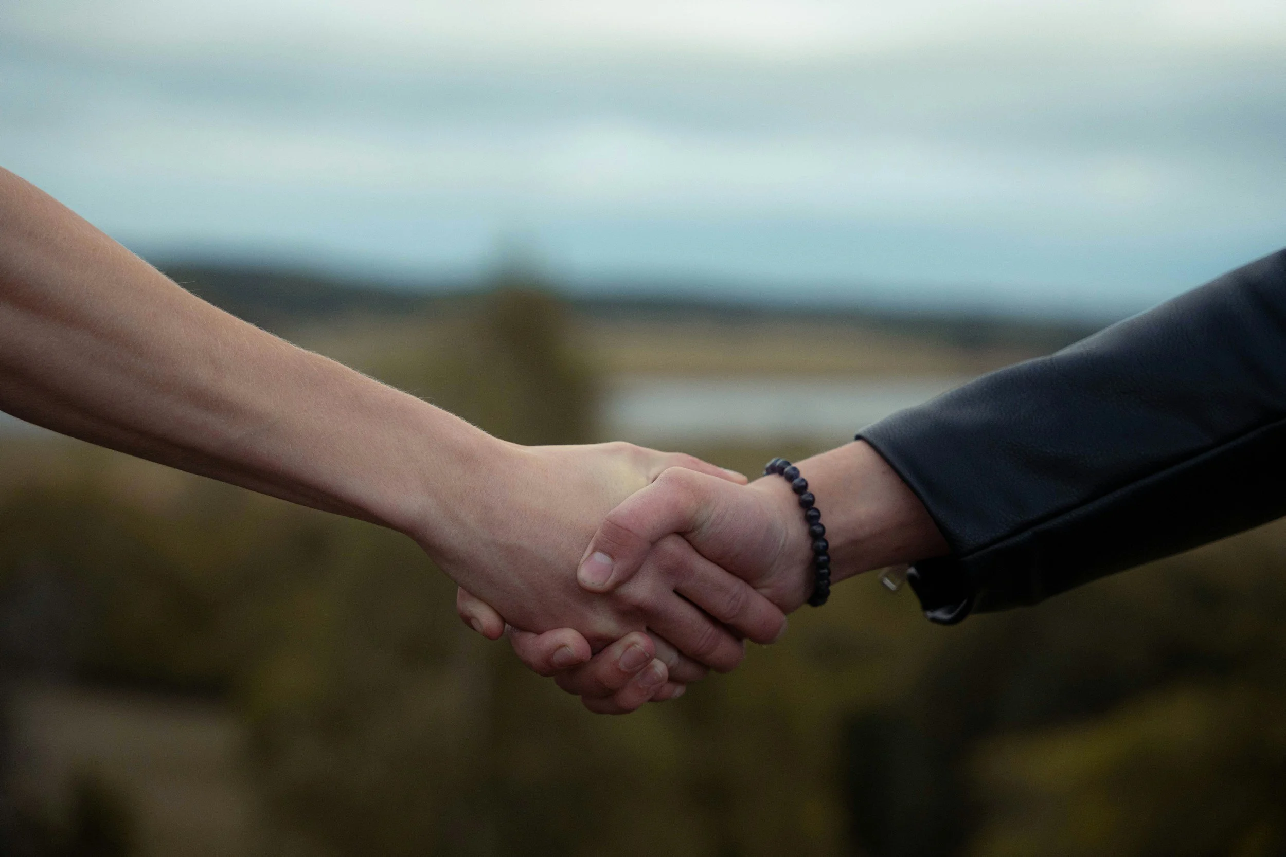 Close-up of two people shaking hands outdoors with a blurred landscape background.