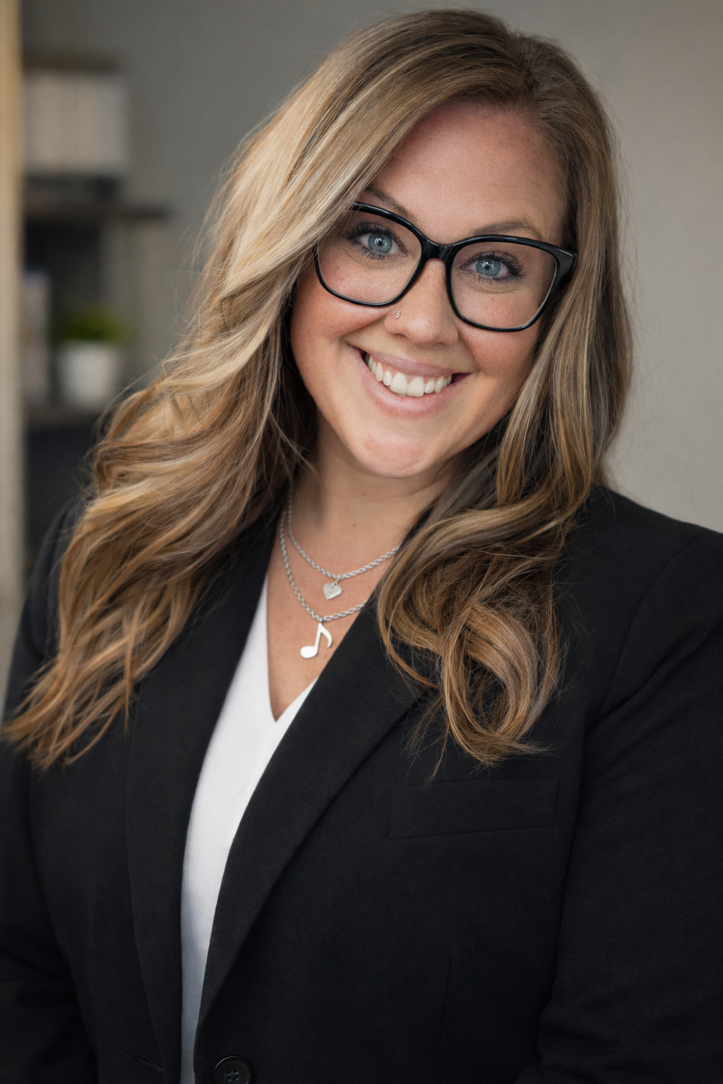 A woman with long, wavy blonde hair, glasses, and a nose piercing smiling in an indoor setting. She wears a black blazer, white top, and layered necklaces, one with a musical note and another with a heart pendant.
