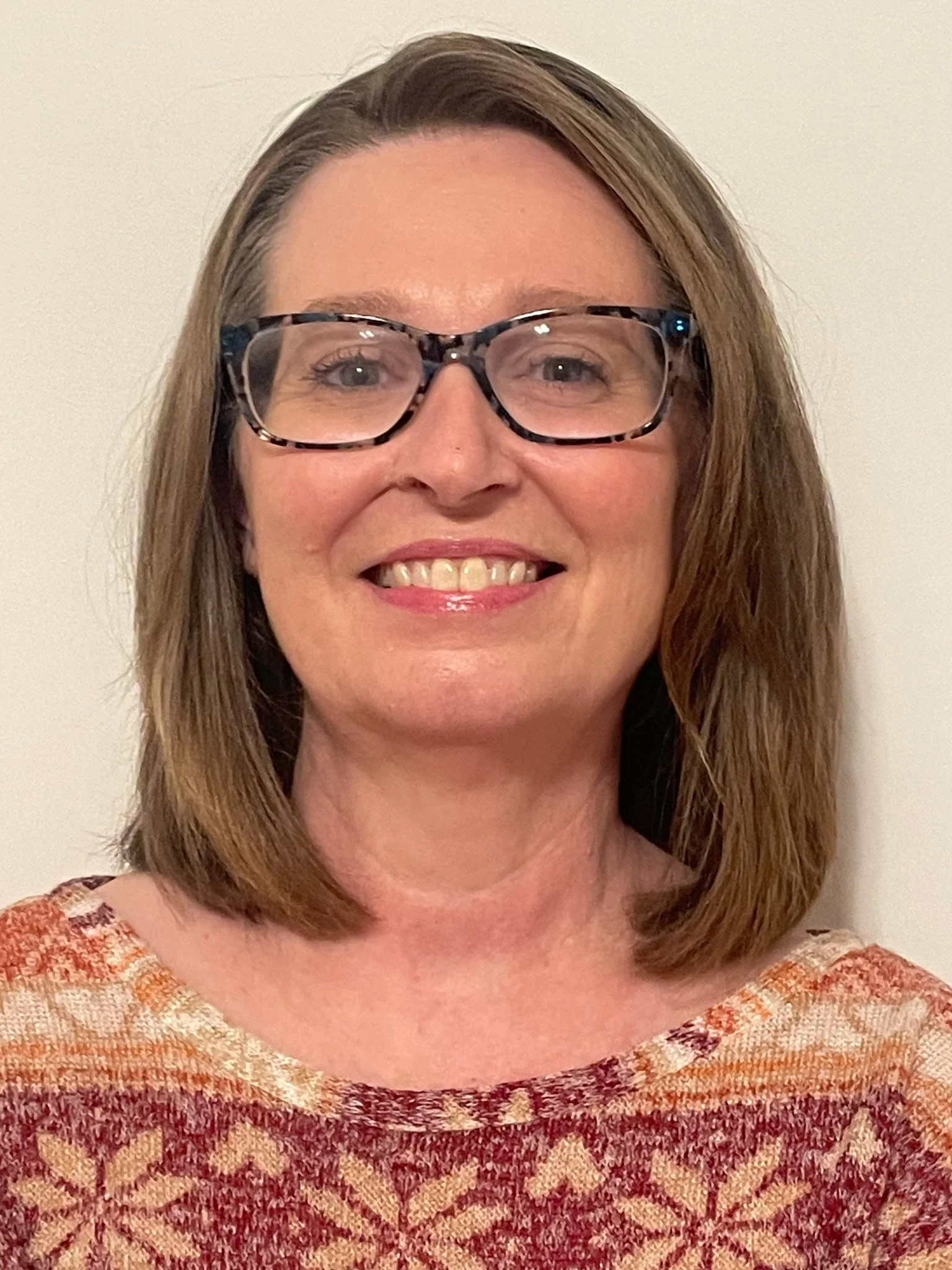 A woman with shoulder-length brown hair, wearing glasses and a patterned sweater, smiling in front of a plain white wall.