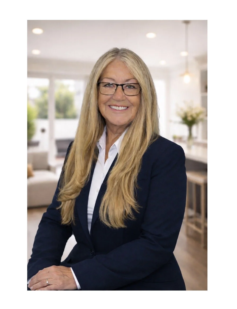 A middle-aged woman with long blonde hair, glasses, and a white shirt under a dark blazer, sitting in a bright, modern room with large windows and a living area in the background.