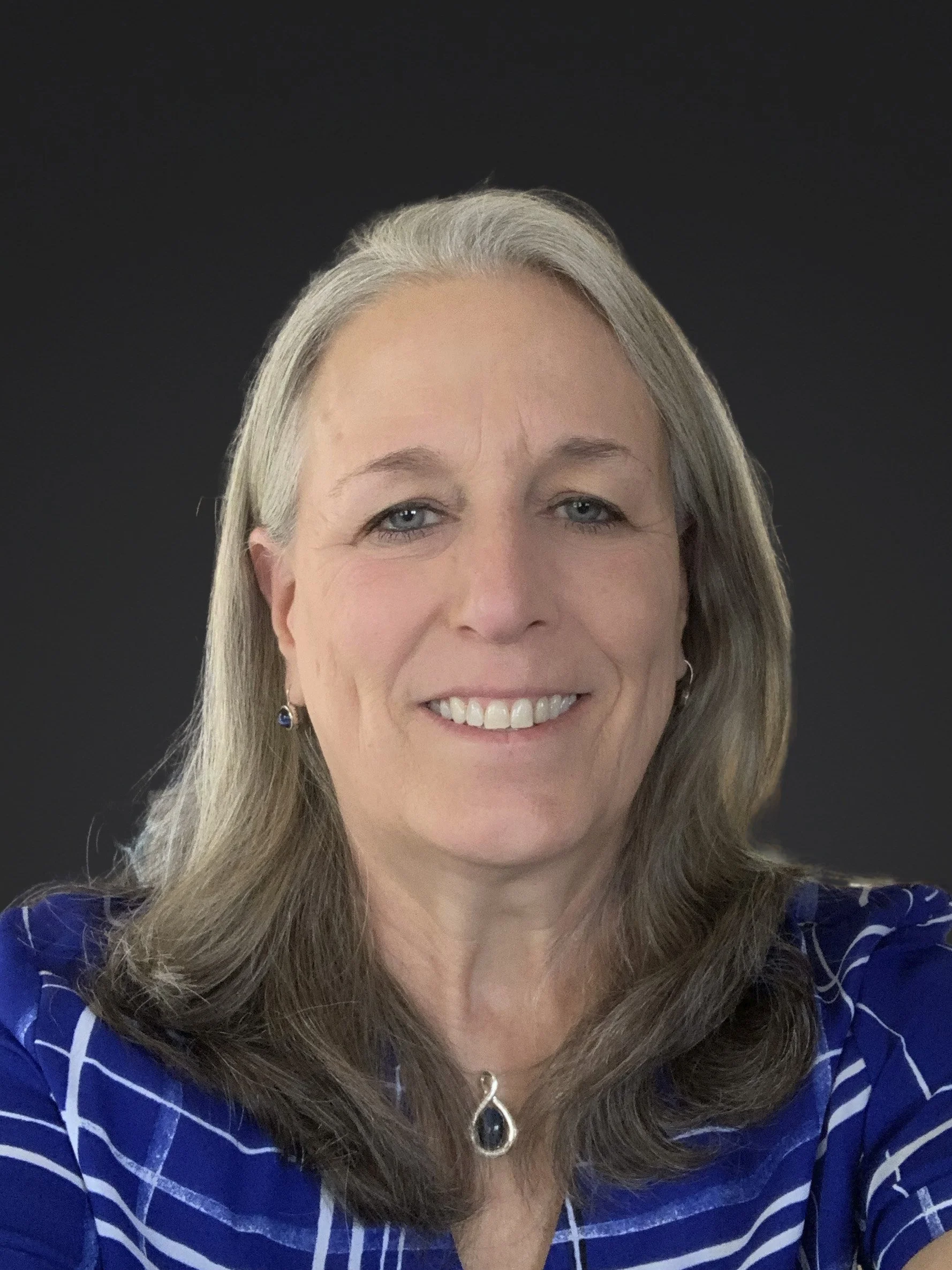 A woman with long gray hair, wearing a blue striped blouse, earrings, and a necklace, smiling against a dark background.
