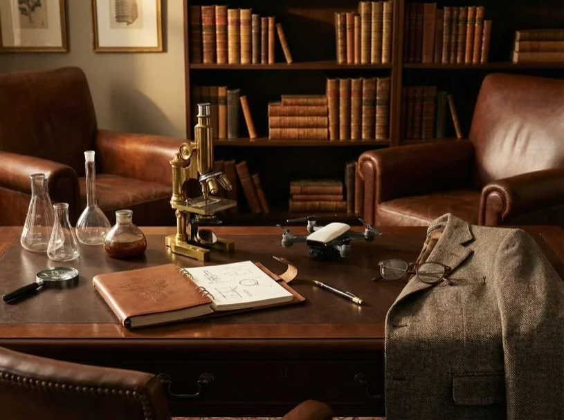 A study room with a wooden desk holding scientific glassware, a notebook, a pen, and a mini drone. There are leather chairs, a gray blazer with glasses on top, and a bookcase filled with old books in the background.