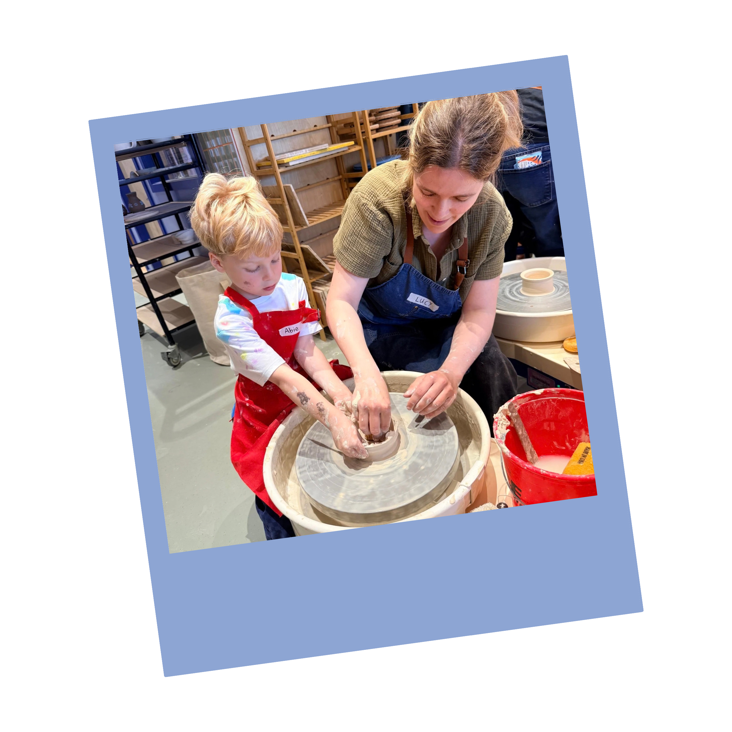 A woman and a young boy are engaged in pottery making at a pottery studio. The boy is wearing a red apron with a name tag that reads 'Abie,' and the woman is wearing a blue apron with a name tag that reads 'Lucy.' They are working together with clay on a pottery wheel, with various pottery tools and a red bucket nearby.