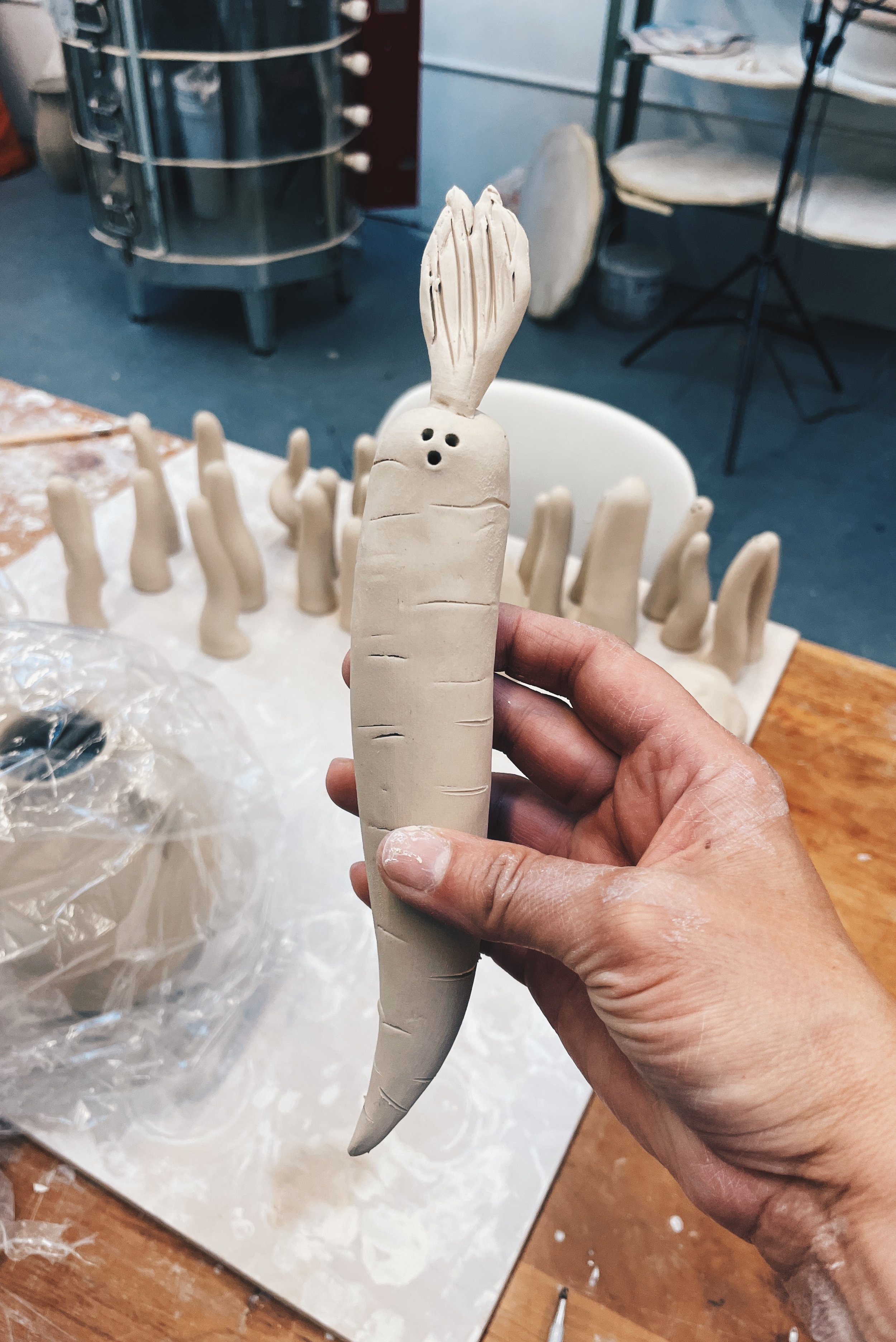 A hand holds up a hand built carrot ghost in front of a plastic covered vase and a bunch of little ghosts made during a workshop