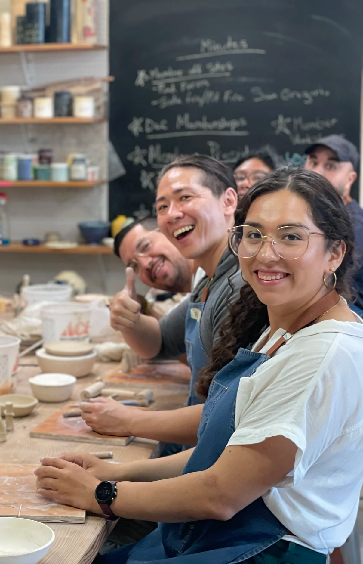 A happy team does hand building together at a long table. One man gives a big thumbs up