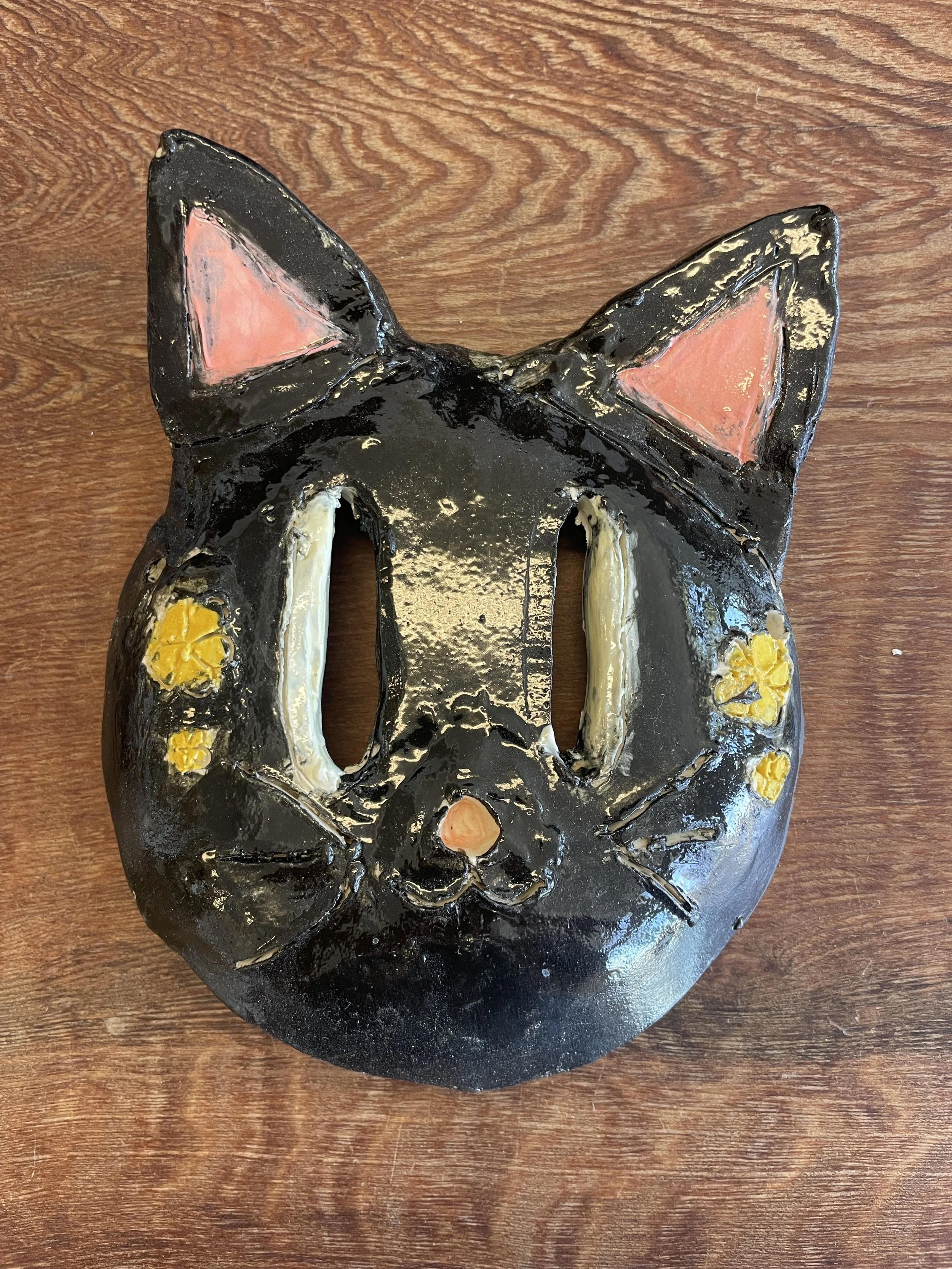 Cat mask made with clay. Black with yellow speckles on its cheeks and pink ears