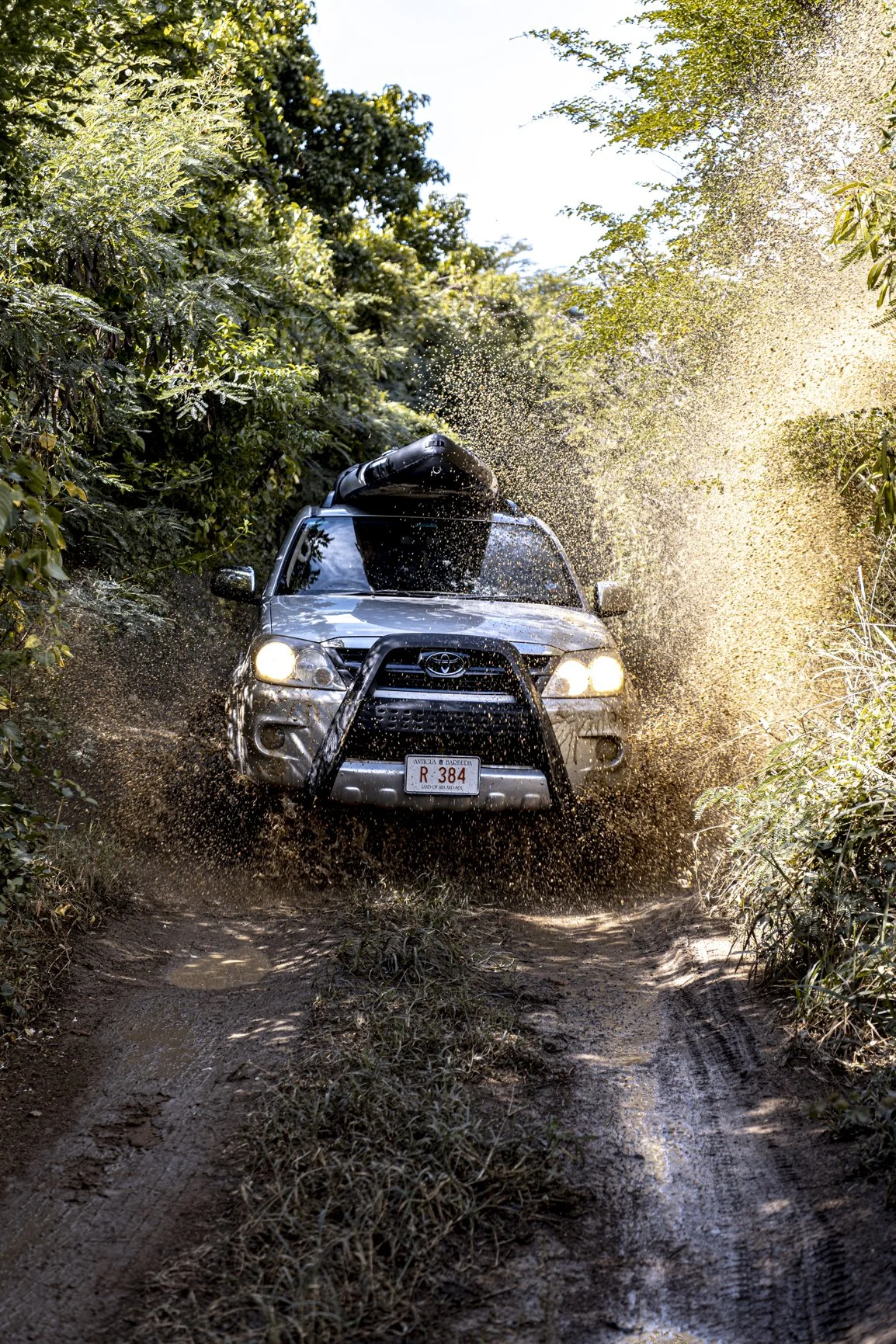 A silver Toyota SUV driving through a muddy off-road trail, splashing dirt and water, surrounded by green trees and bushes.