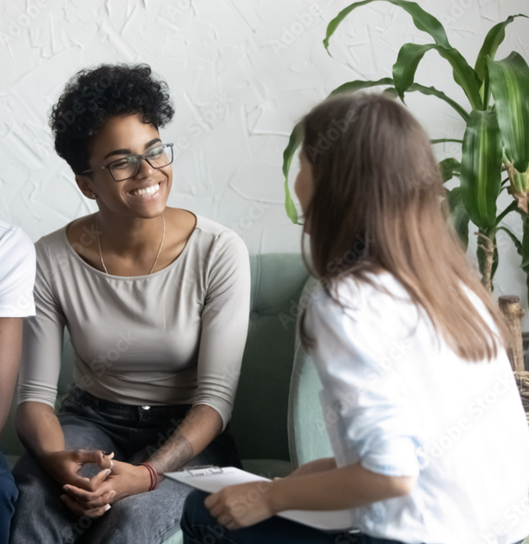 Two women having a conversation in a cozy room with a green plant in the background. One woman is smiling and wearing glasses, the other has long hair and holds a clipboard.