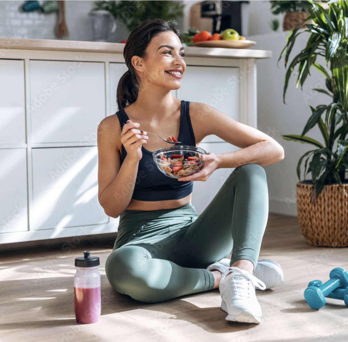 A woman in workout clothes sitting on the floor in a bright kitchen, eating healthy cereal with fresh berries from a glass bowl.